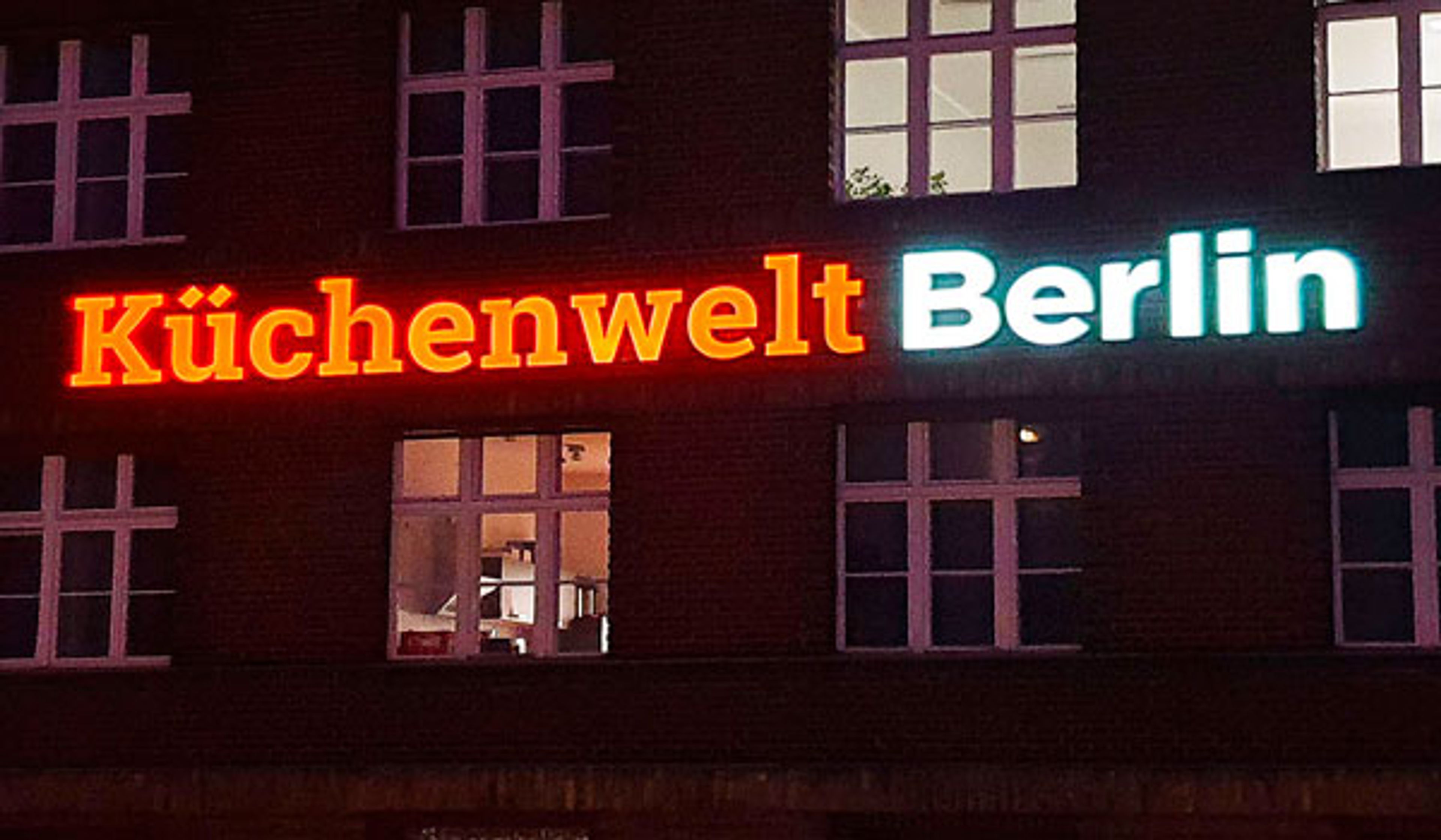 Orange und weiß leuchtender Schriftzug „Küchenwelt Berlin“ an einer Backsteinfassade bei Nacht, Nahansicht