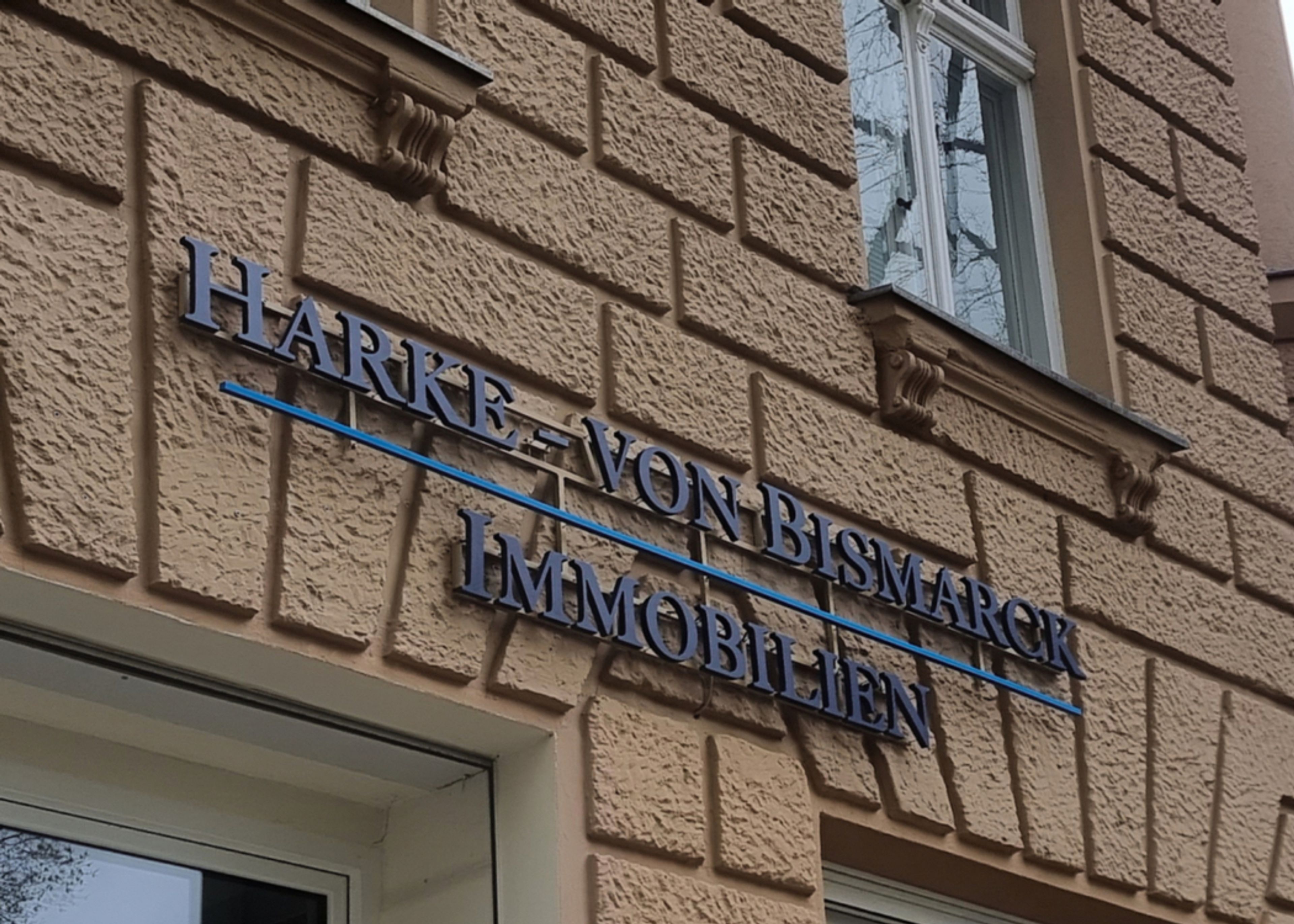 Zweizeiliger Schriftzug aus dunkelblauen Buchstaben mit blauer Linie an einer Putzfassade, von schräg unten