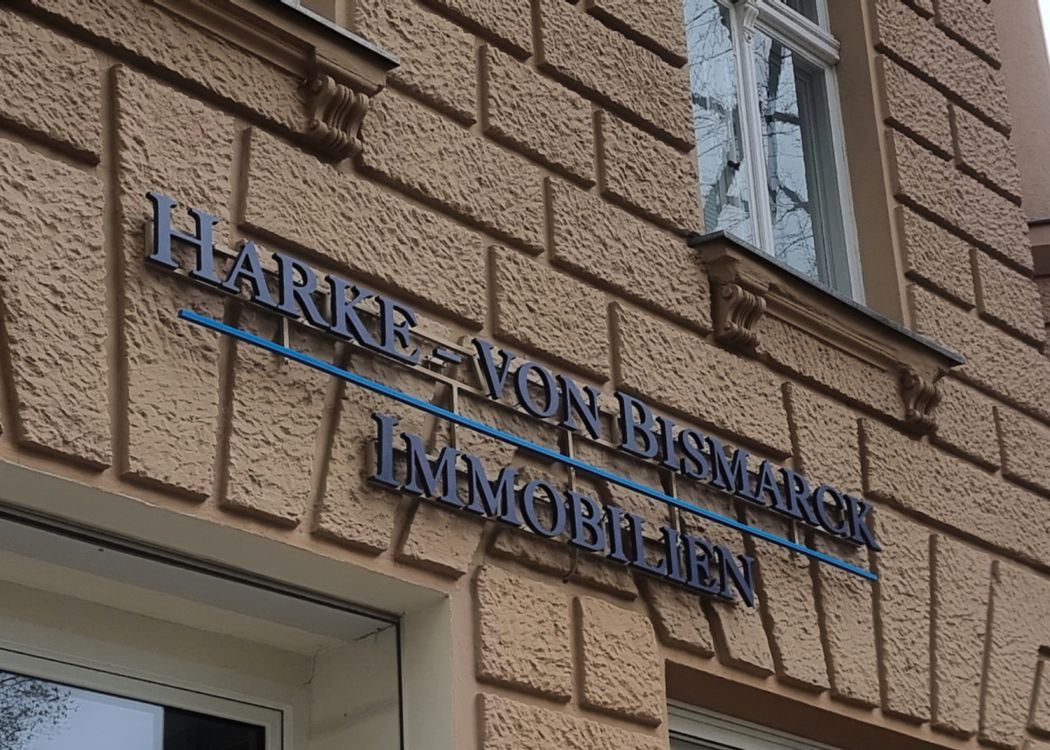 Zweizeiliger Schriftzug aus dunkelblauen Buchstaben mit blauer Linie an einer Putzfassade, von schräg unten