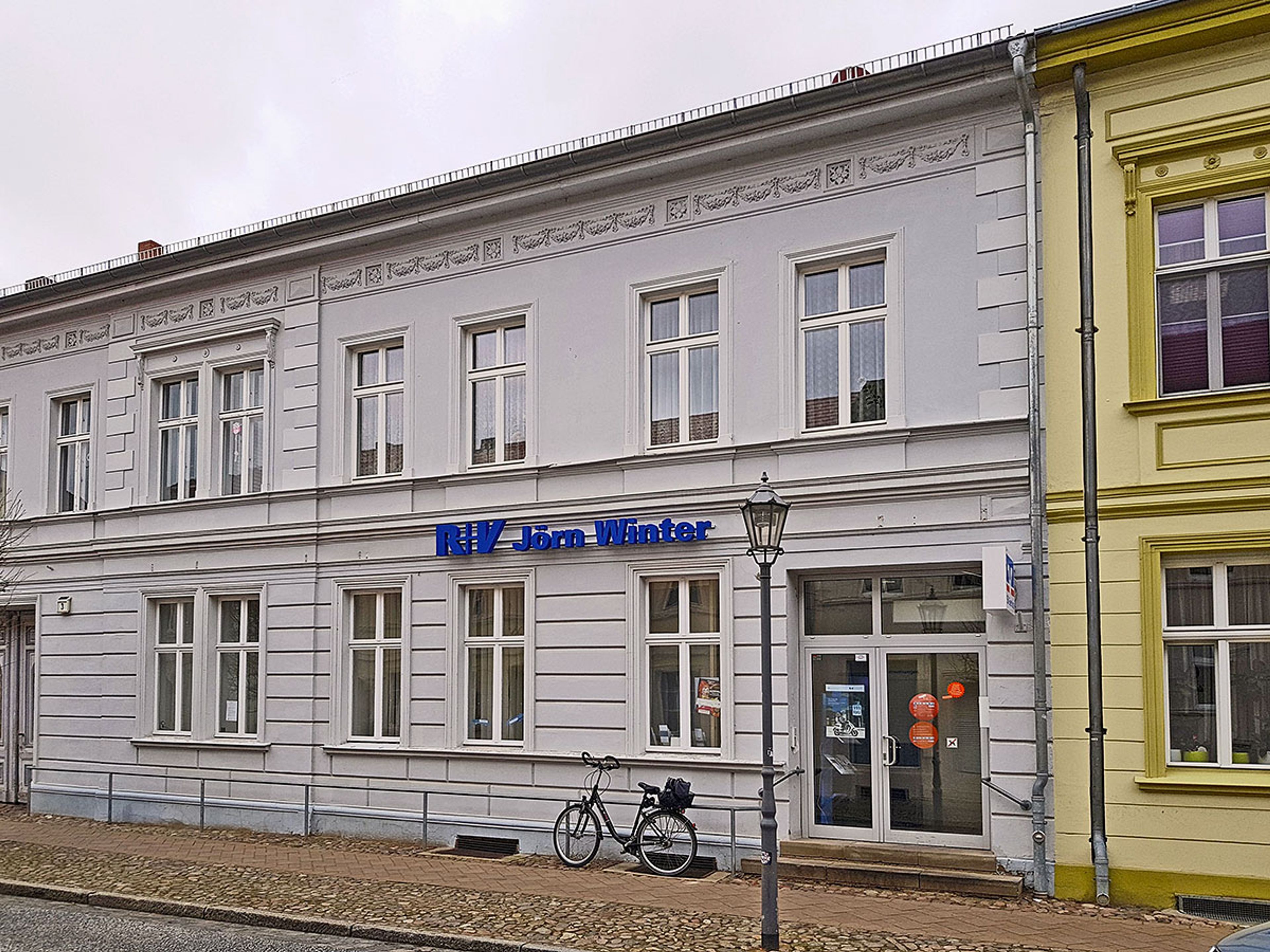 Zweistöckiges Altbauhaus bei Tag mit blauem Schriftzug über dem Erdgeschoss, davor ein Fahrrad und eine Laterne