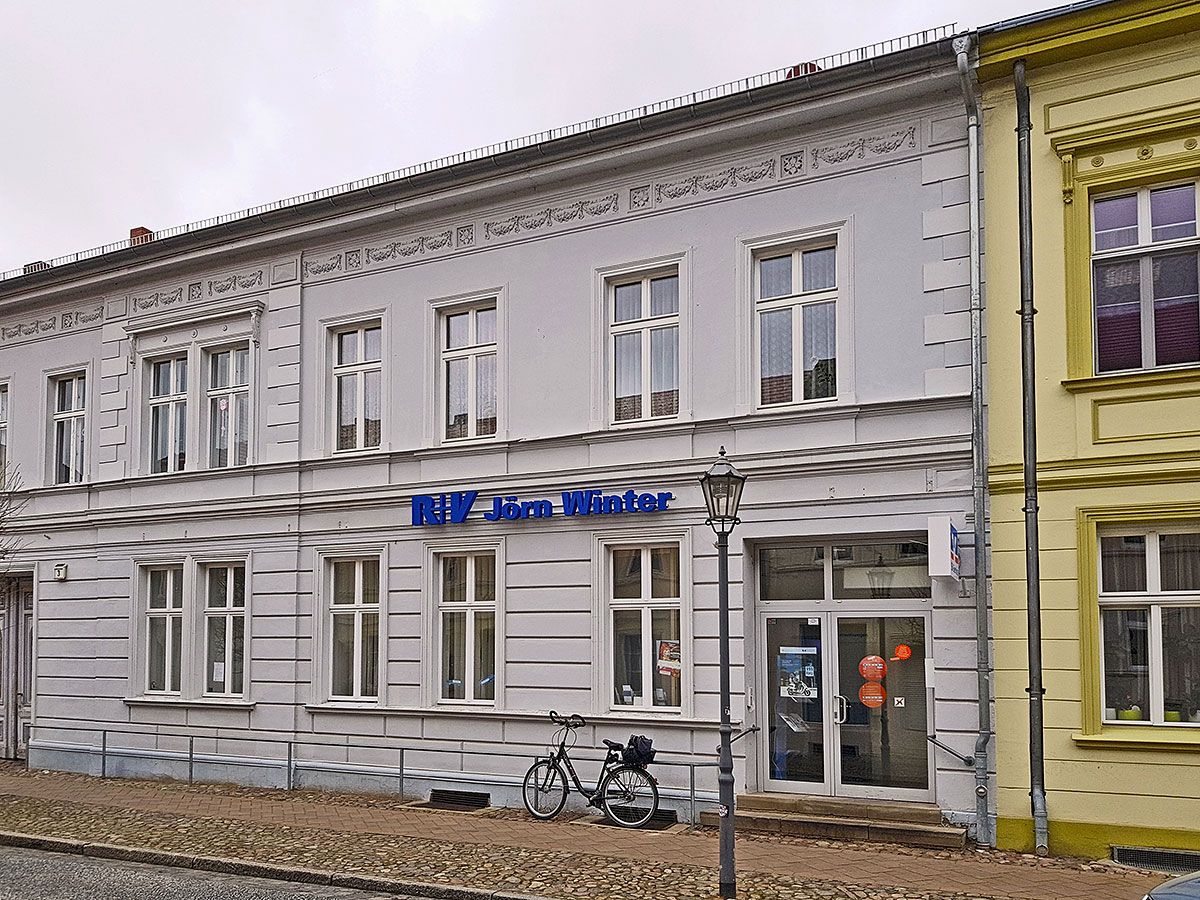 Zweistöckiges Altbauhaus bei Tag mit blauem Schriftzug über dem Erdgeschoss, davor ein Fahrrad und eine Laterne