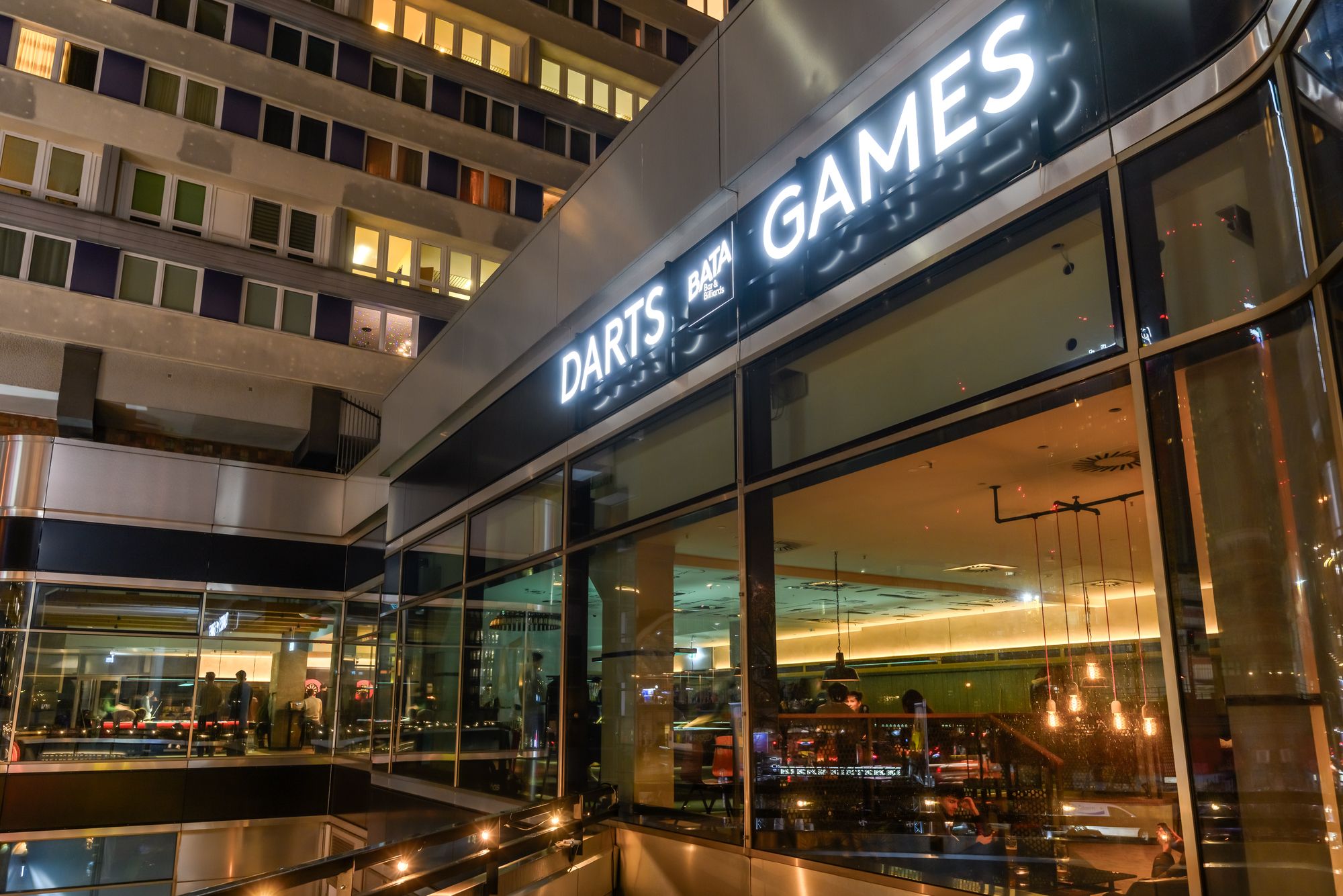 Leuchtende Schriftzüge „Darts“ und „Games“ mit Logo über der Glasfront einer Bar am Abend, von schräg unten