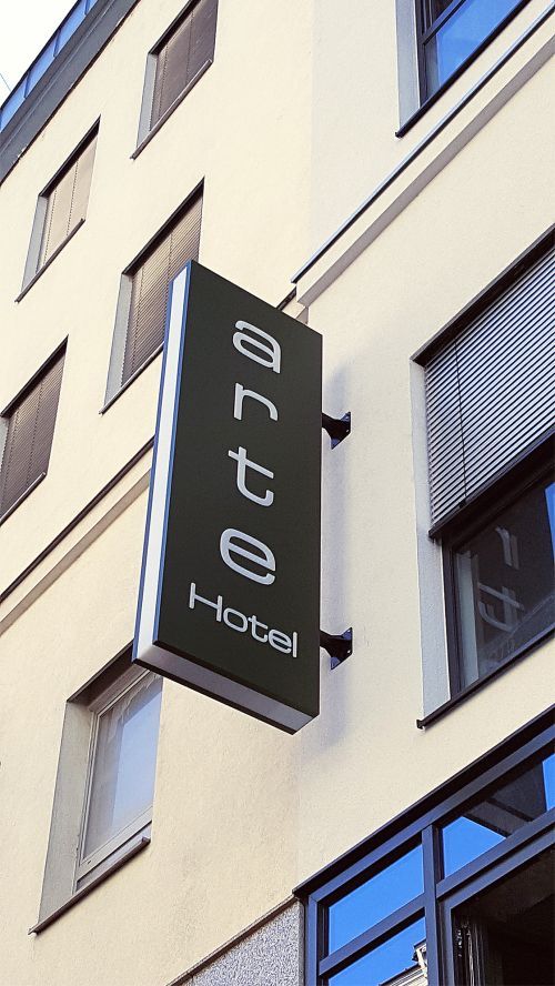 Senkrechter Leuchtkasten quer zur Fassade eines Hotels, bei Tag