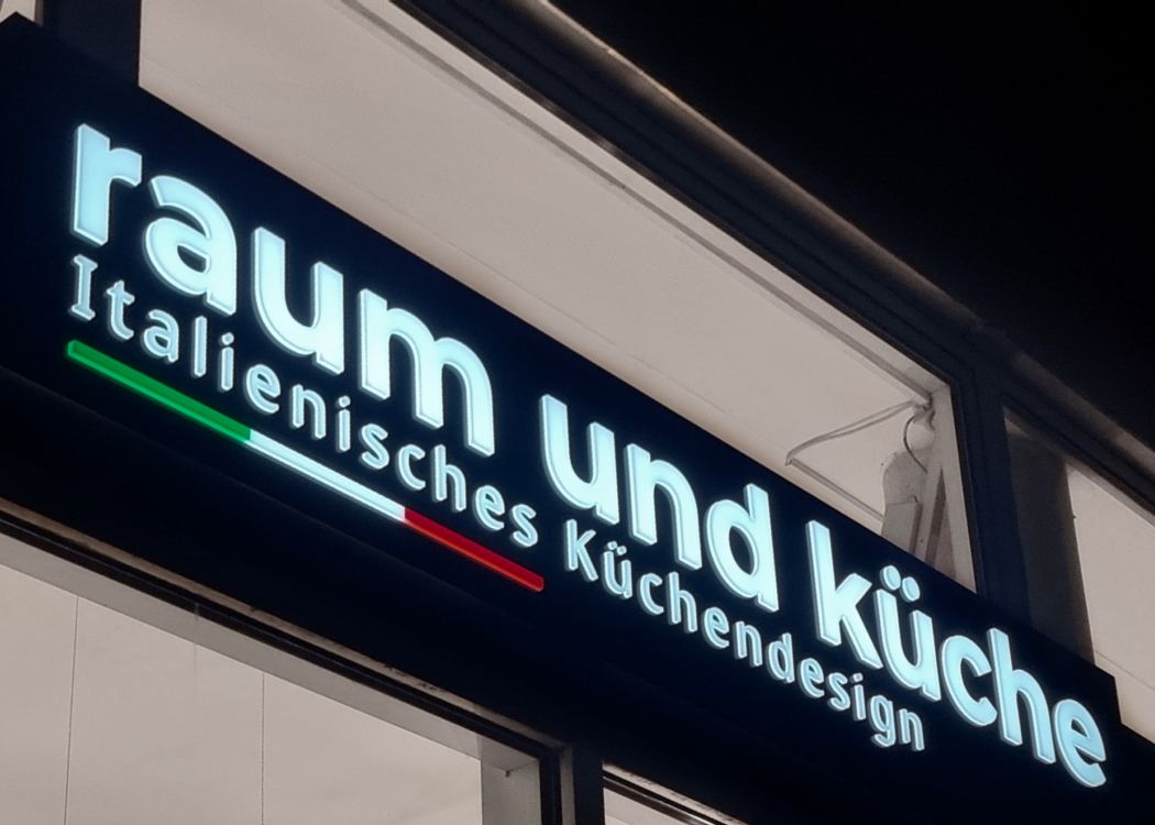 Leuchtkasten mit leuchtender, erhabener Schrift „Raum und Küche“ über einem Schaufenster am Abend, von schräg unten