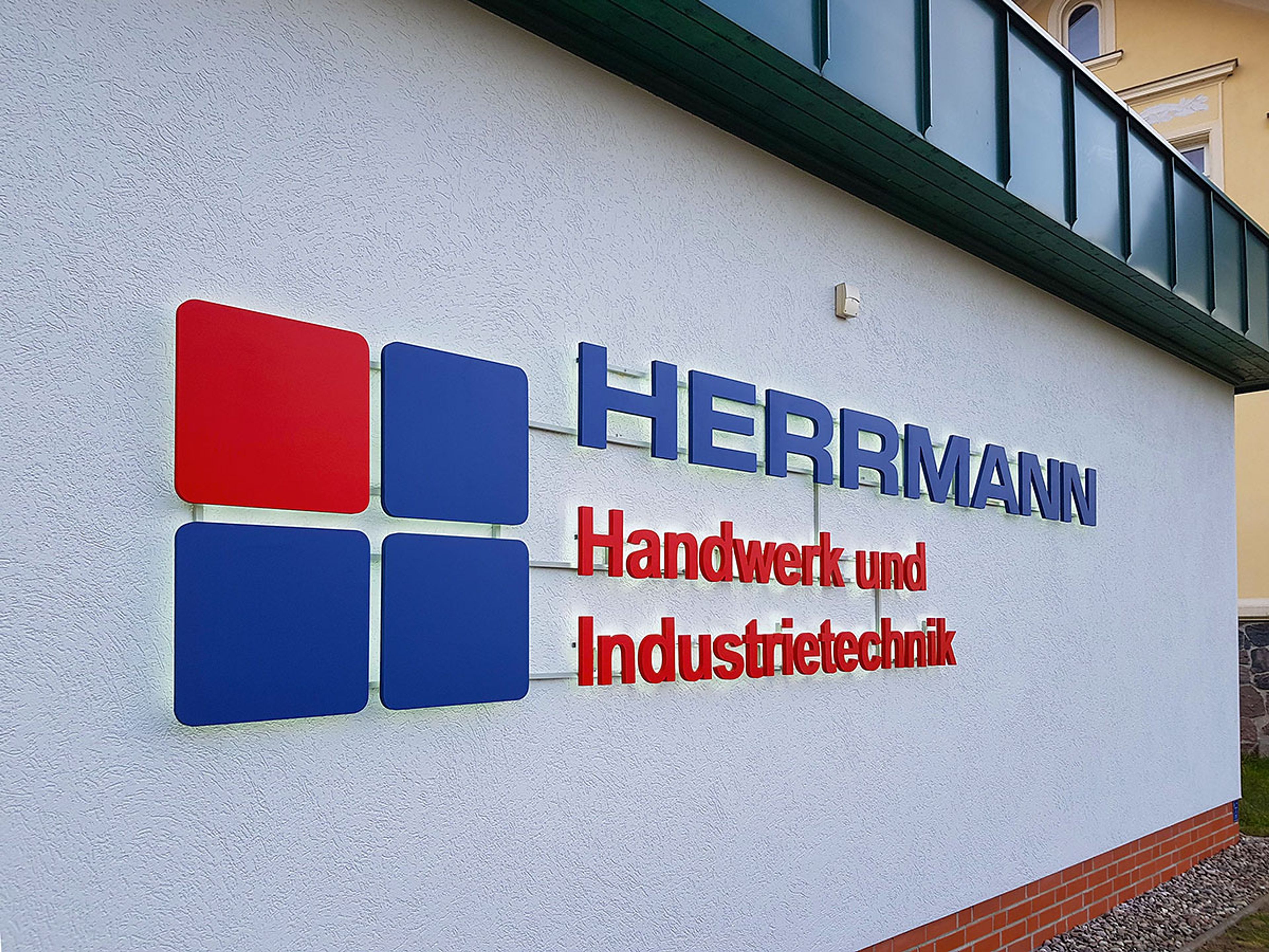 Logo aus roten und blauen Quadraten mit Schriftzug auf Abstandshaltern an einer weißen Putzwand, bei Tag