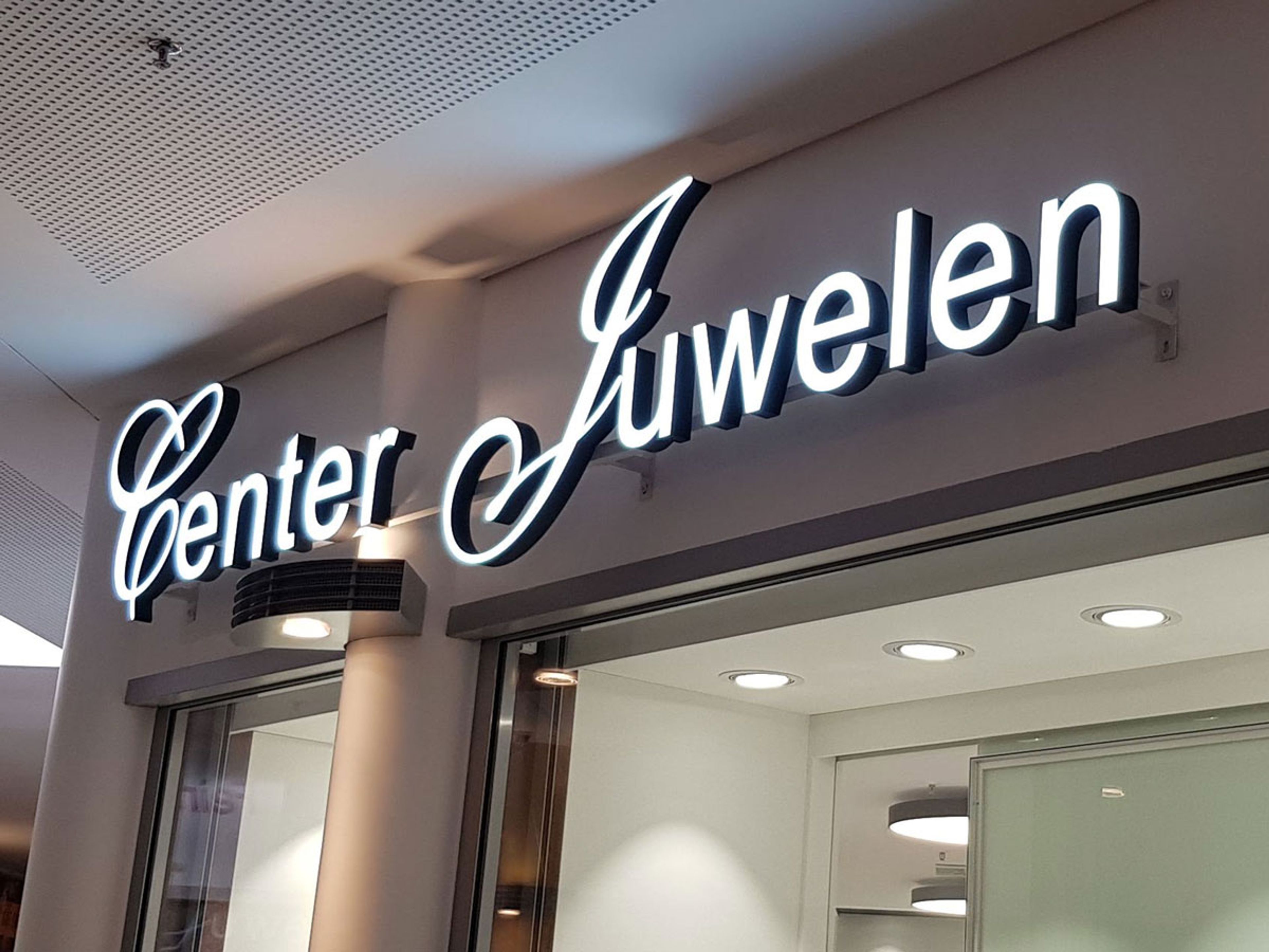 Leuchtender Schriftzug „Center Juwelen“ in Schreibschrift über einem Ladeneingang im Innenbereich, von schräg unten