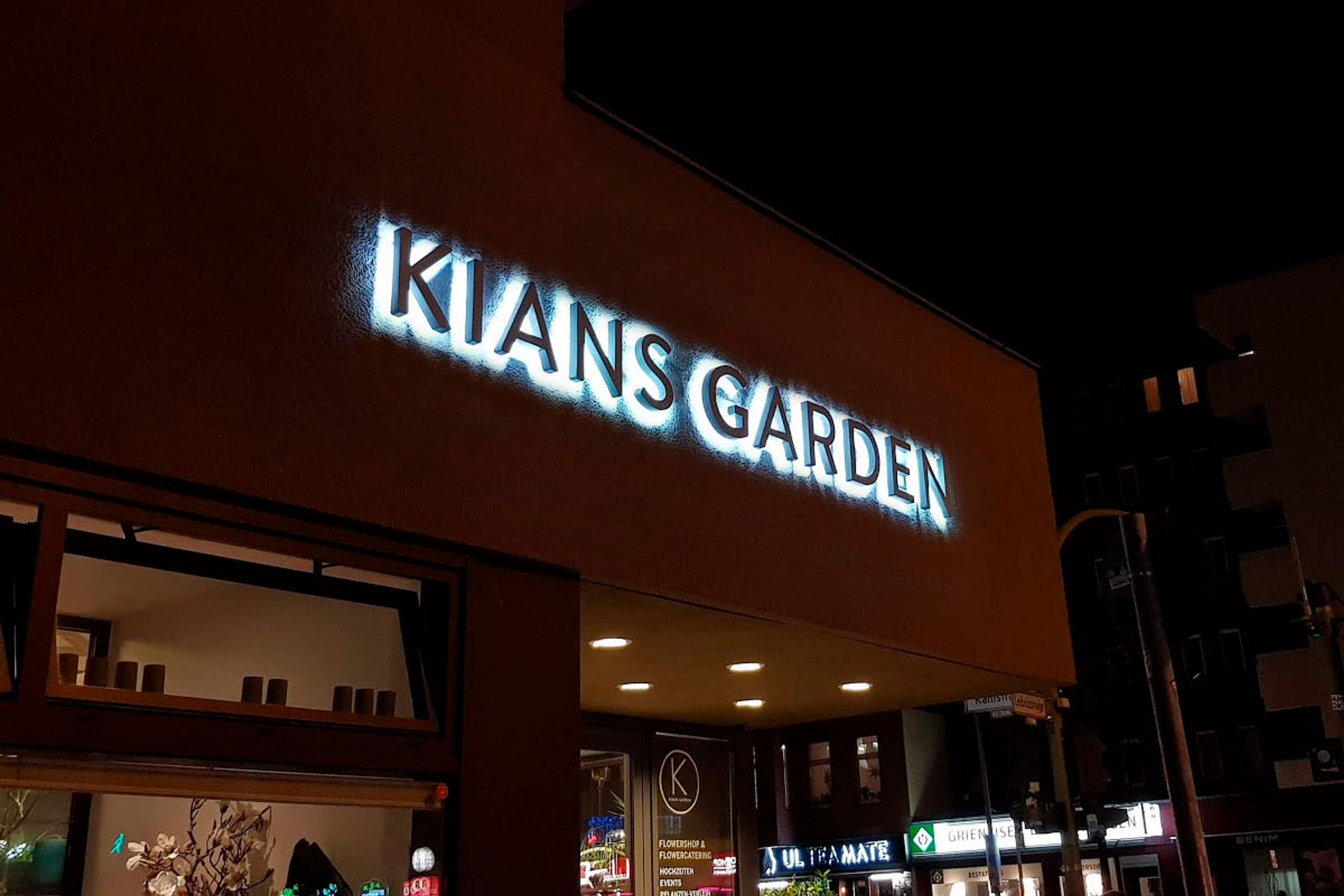 Weiße Buchstaben mit hellem Lichtschein an einer dunklen Fassade über einem Ladeneingang, bei Nacht