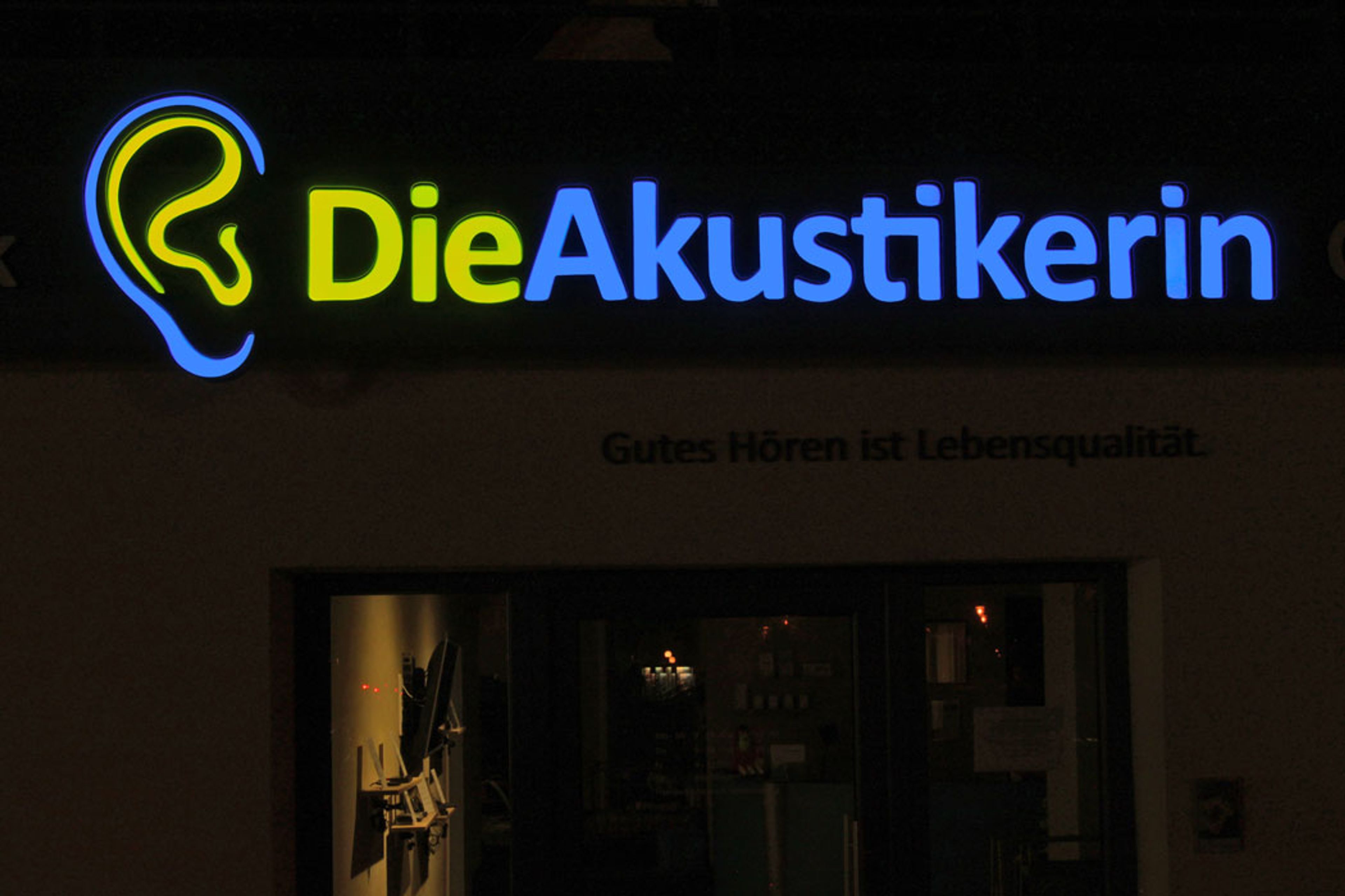 Nahaufnahme des leuchtenden Ohr-Logos in Gelb und Blau und des Schriftzugs „Die Akustikerin“ bei Nacht
