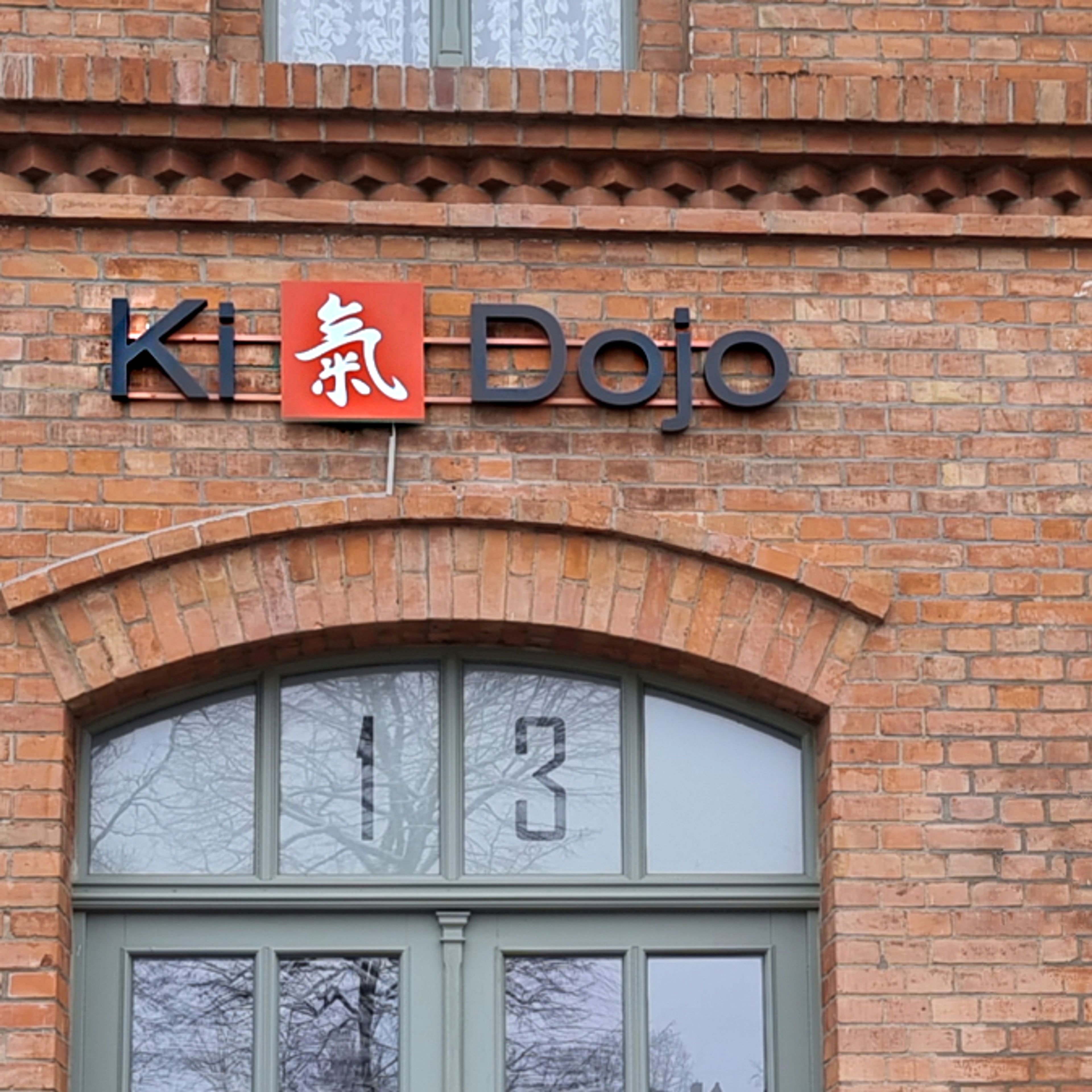 Buchstaben mit rotem Logo an einer Backsteinfassade über einem Rundbogenfenster