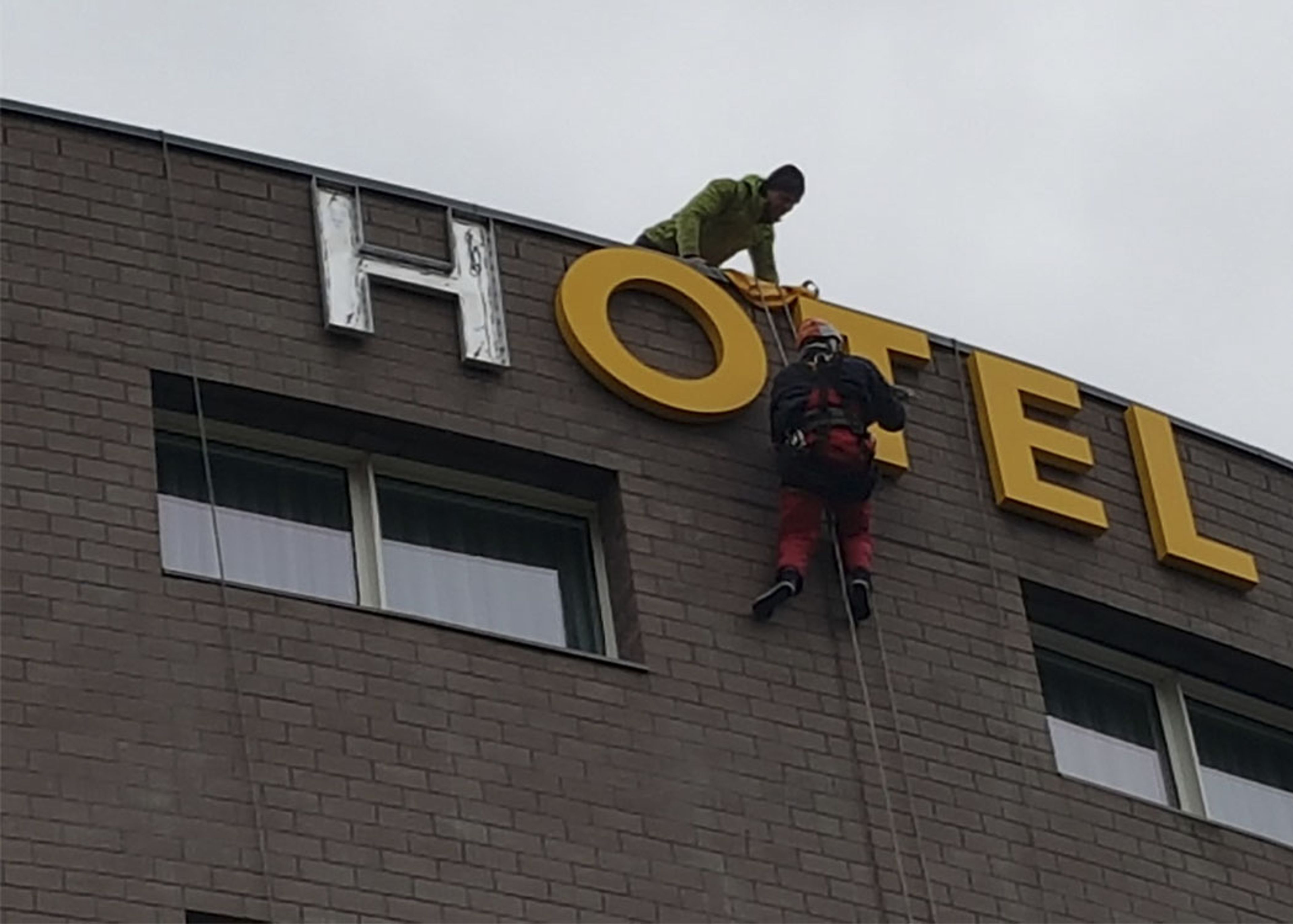 Monteure am Seil bringen große gelbe Buchstaben oben an der Klinkerfassade eines Hotels an, links ein alter Buchstabe