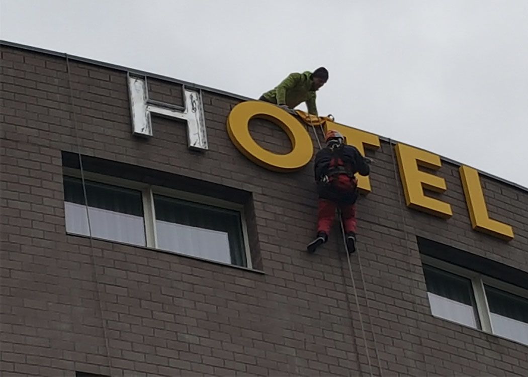 Monteure am Seil bringen große gelbe Buchstaben oben an der Klinkerfassade eines Hotels an, links ein alter Buchstabe