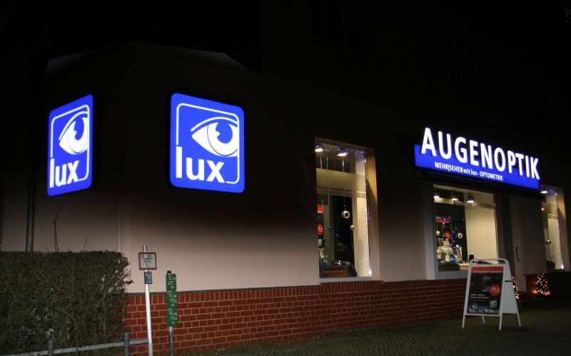 Augenoptik-Geschäft am Abend mit beleuchtetem Logo-Kasten und Leuchtschrift über dem Schaufenster