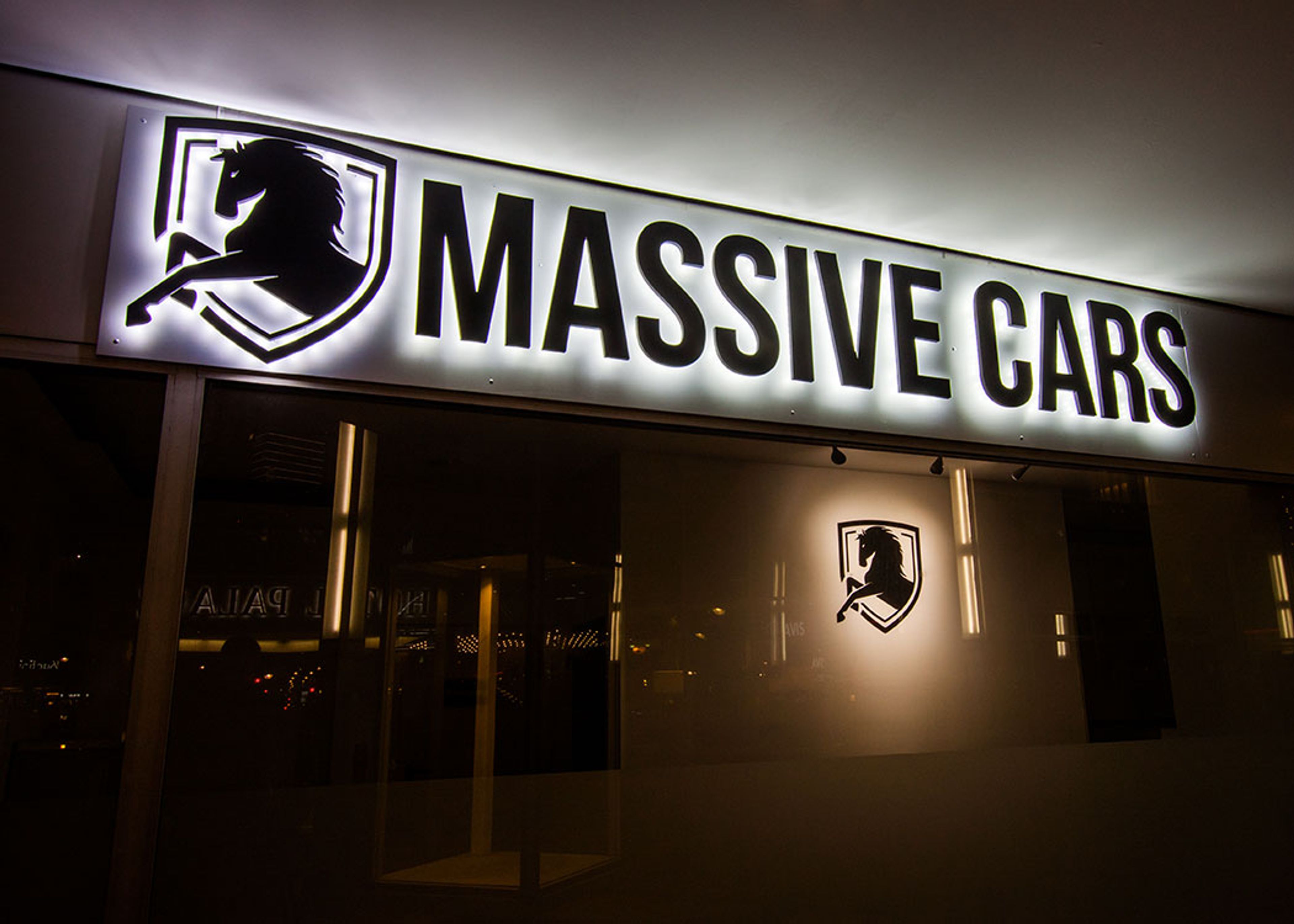 Nahaufnahme von „Massive Cars“ mit Pferdelogo und hellem Lichtschein, im Schaufenster darunter ein weiteres Logo