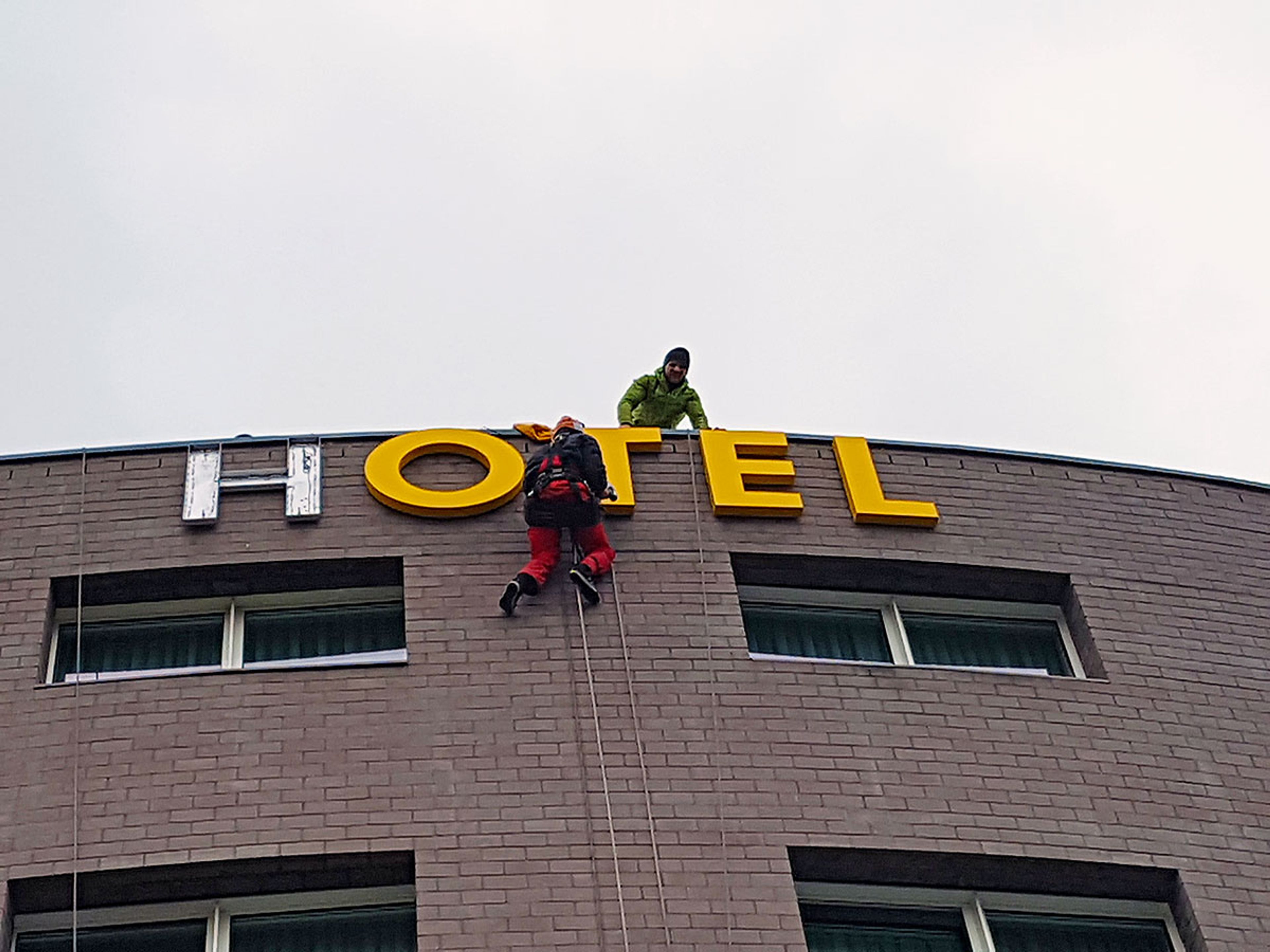 Zwei Monteure, einer am Seil hängend, setzen die neuen gelben Buchstaben „Hotel“ an der Dachkante ein