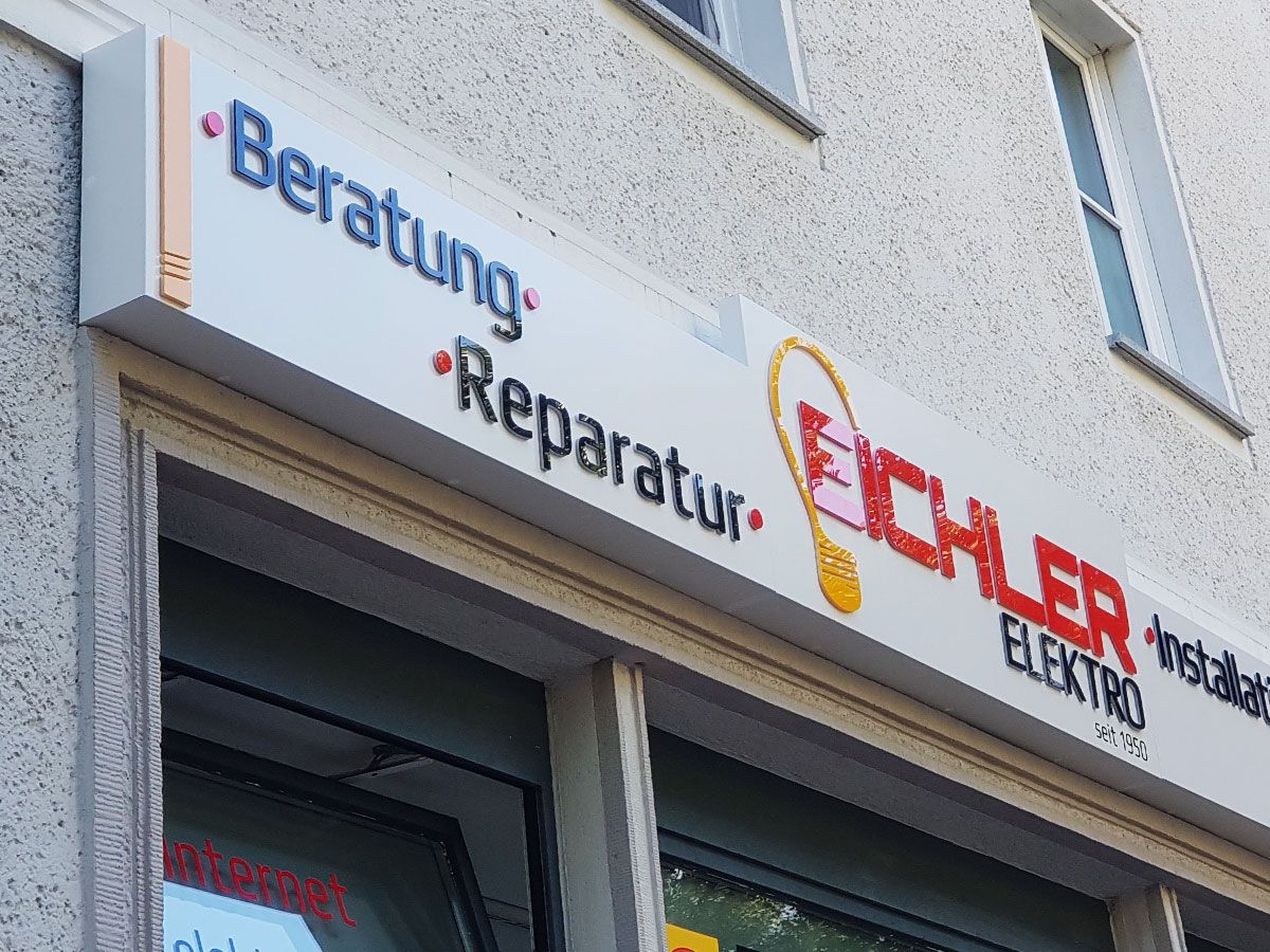 Weißer Leuchtkasten mit erhabenen Schriftzügen und Glühbirnen-Logo über einem Ladengeschäft, von schräg unten bei Tag