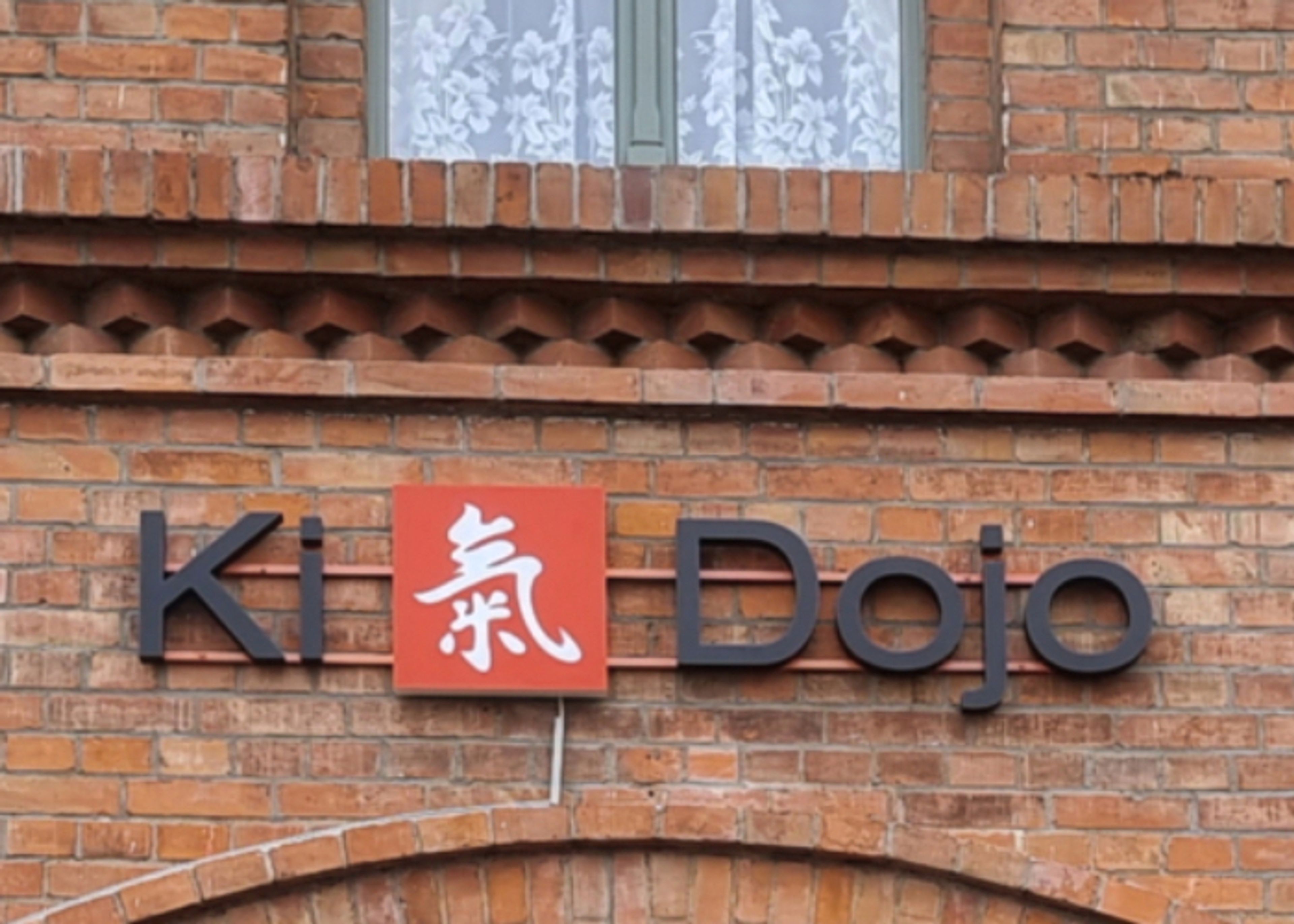 Nahaufnahme der dunklen Buchstaben „Ki Dojo“ und des roten Logofelds auf Abstandshaltern an der Backsteinwand