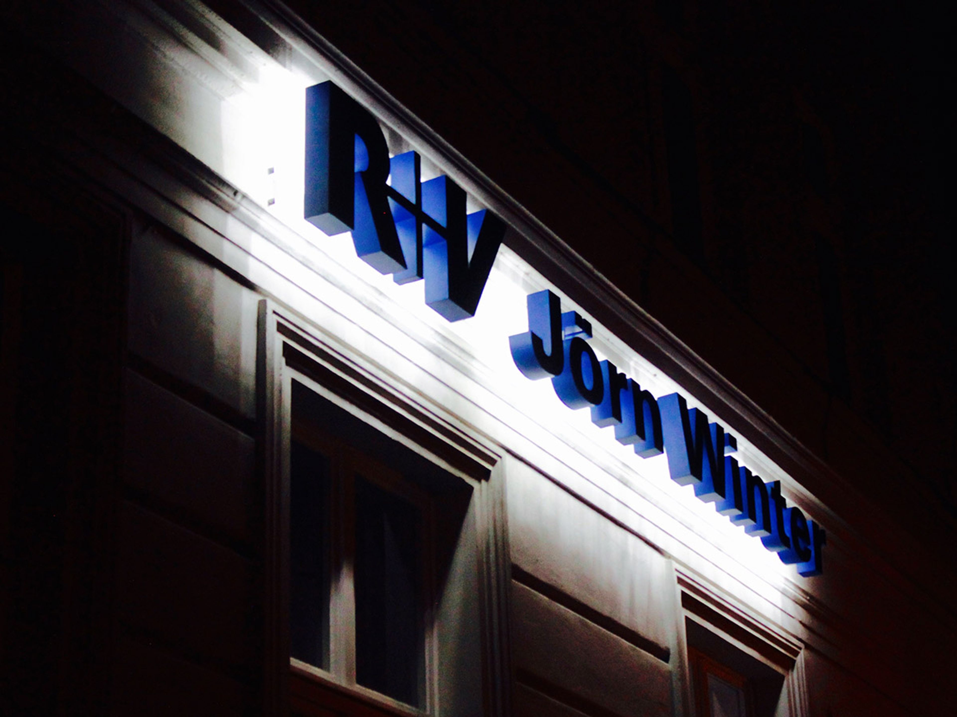 Blaue Buchstaben mit hellem Lichtschein an einer Altbaufassade bei Nacht, von schräg unten