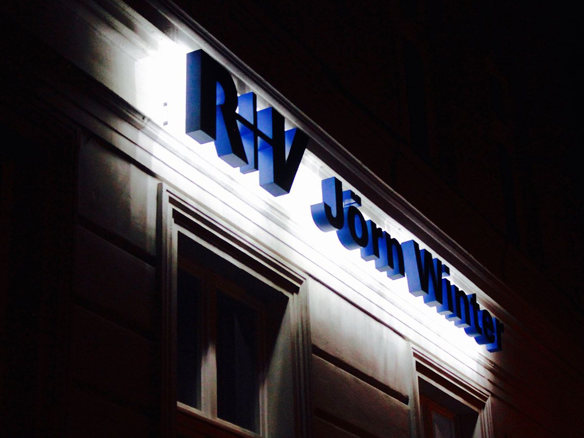 Blaue Buchstaben mit hellem Lichtschein an einer Altbaufassade bei Nacht, von schräg unten