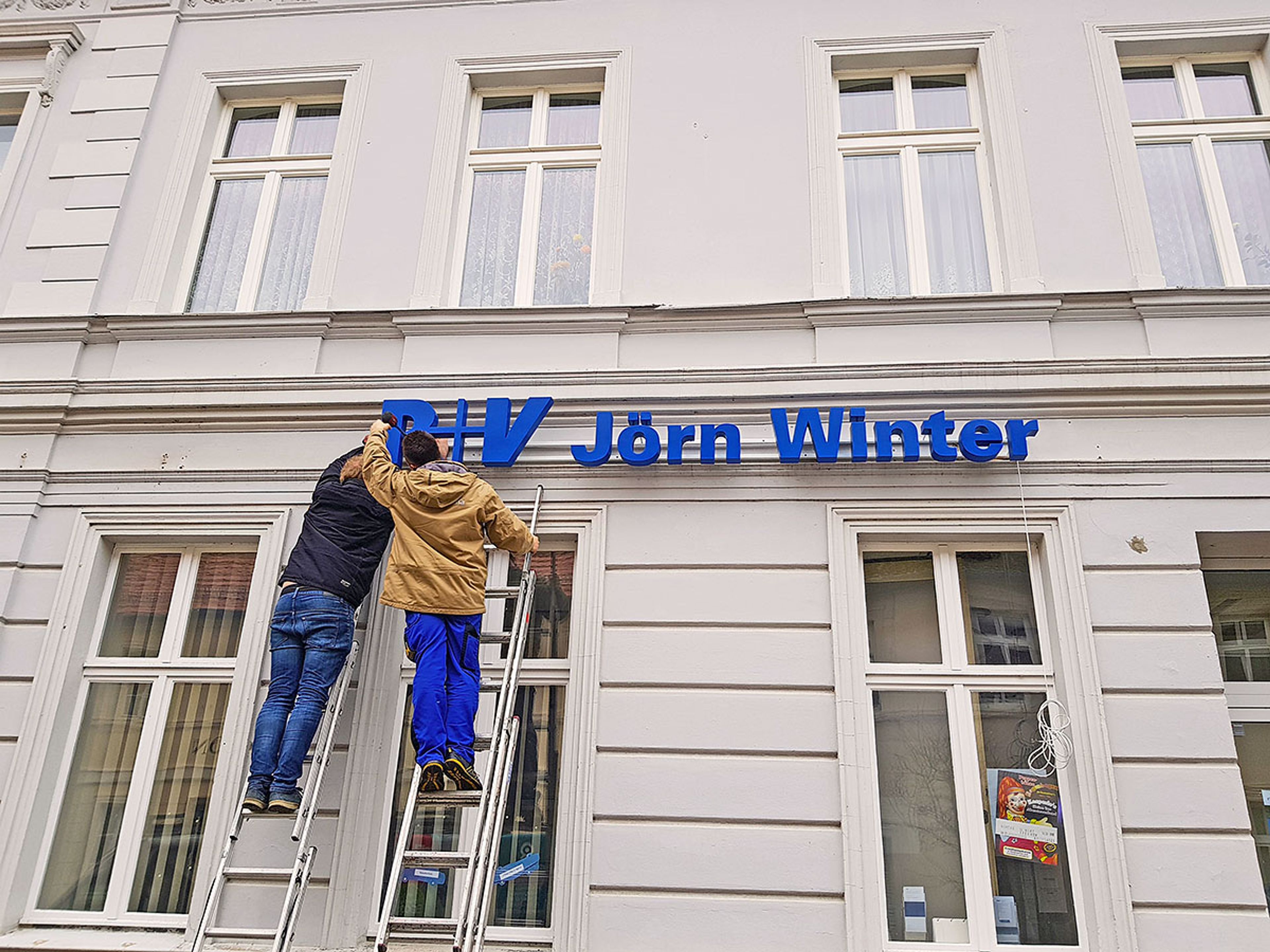 Zwei Monteure auf Leitern bringen blaue Einzelbuchstaben an der Fassade eines Versicherungsbüros an