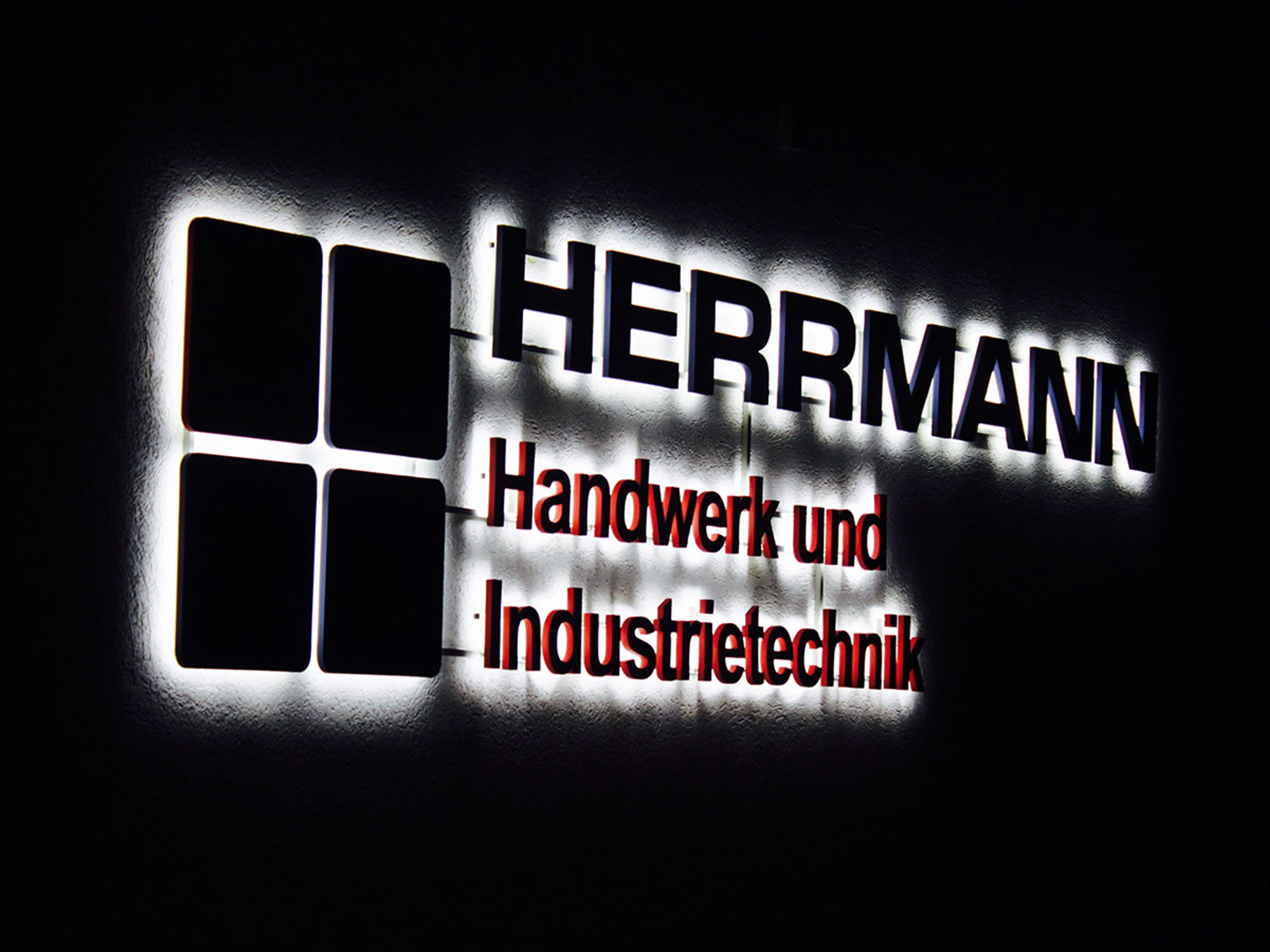 Logo und Schriftzug als dunkle Silhouetten, rundum von hellem Lichtschein an der Wand umgeben, bei Nacht