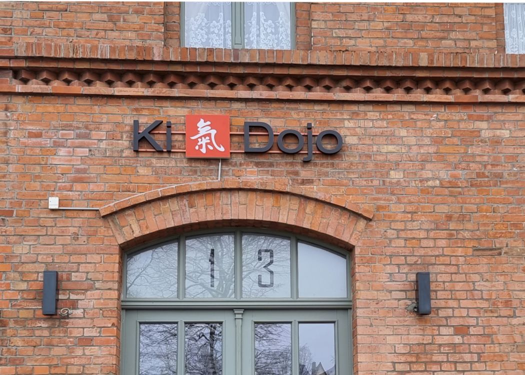 Schriftzug „Ki Dojo“ aus dunklen Buchstaben mit rotem Logofeld über dem Rundbogeneingang eines Backsteinhauses