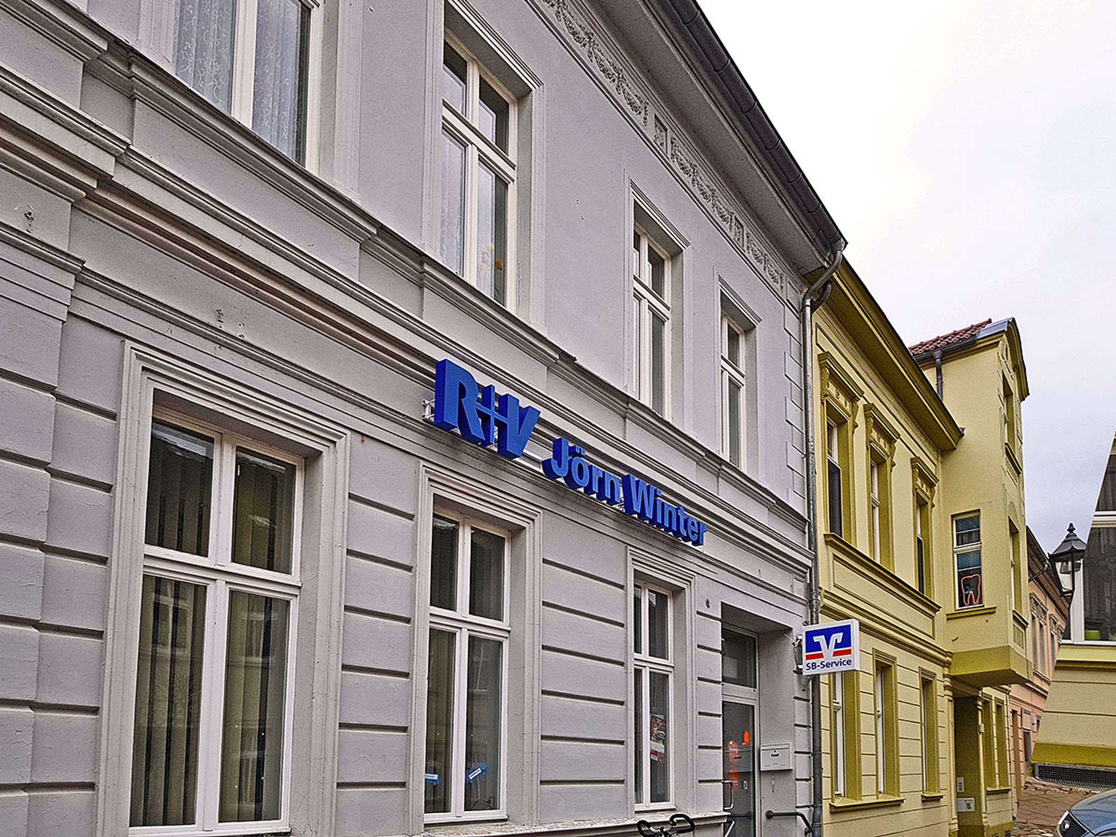 Blaue Buchstaben eines Versicherungsbüros an einer hellen Altbaufassade in einer schmalen Straße, von schräg unten