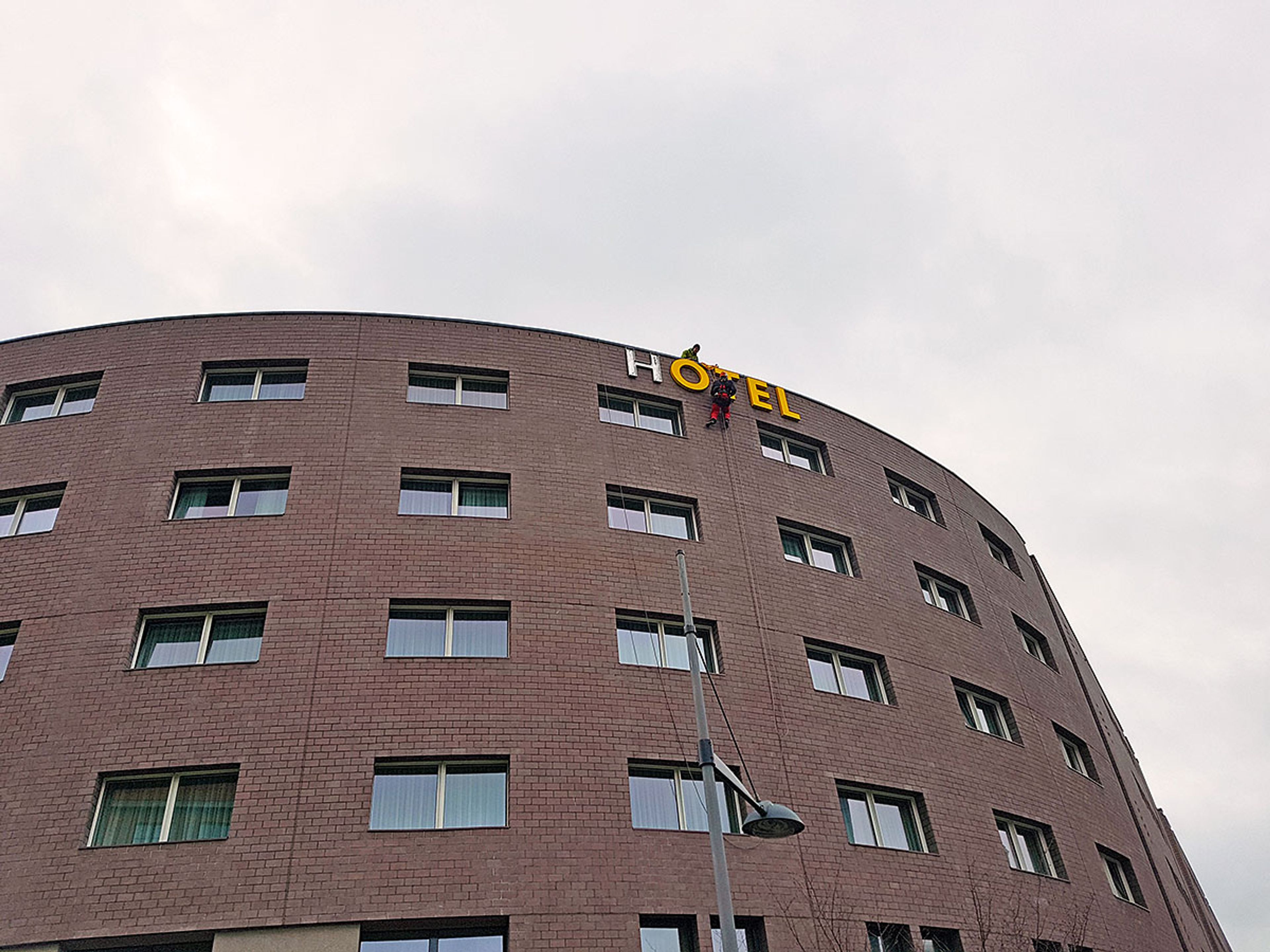 Gebogene Klinkerfassade eines Hotels bei Tag, oben bringen Monteure am Seil die Buchstaben „Hotel“ an