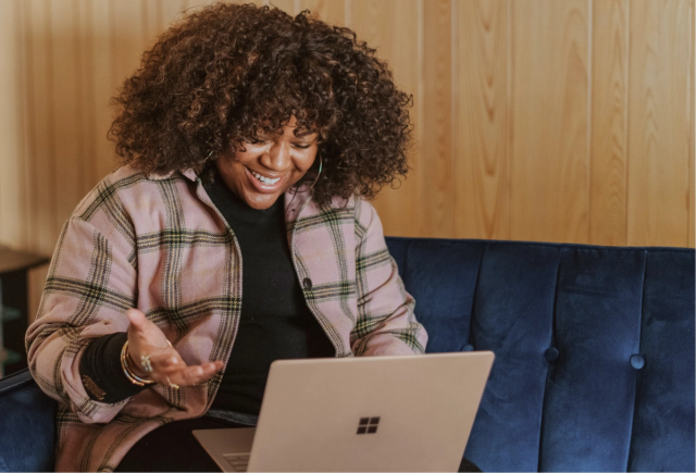 A smiling black woman sits on a blue couch using her laptop