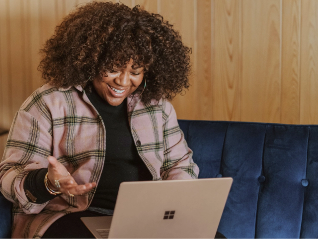 A smiling black woman sits on a blue couch using her laptop