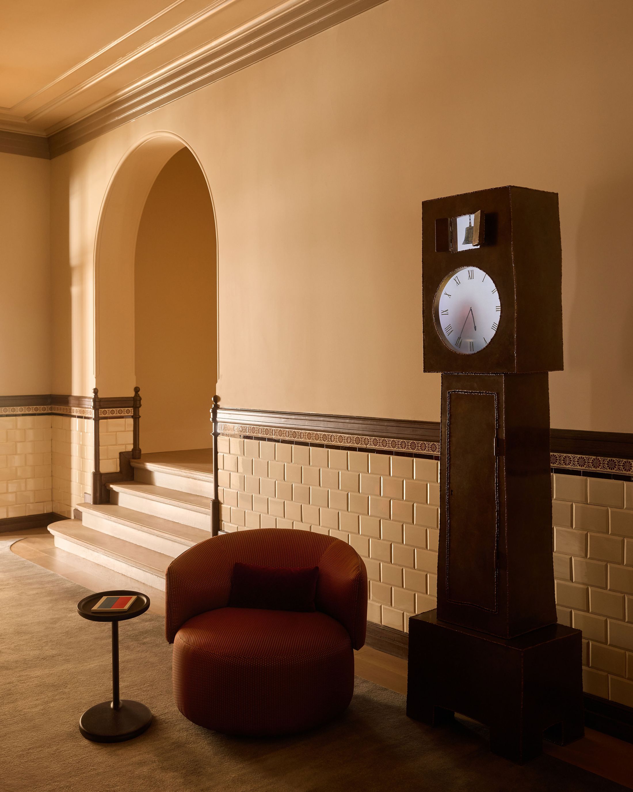 A grandfather clock next to a red leather chair and table in a minimalist corridor