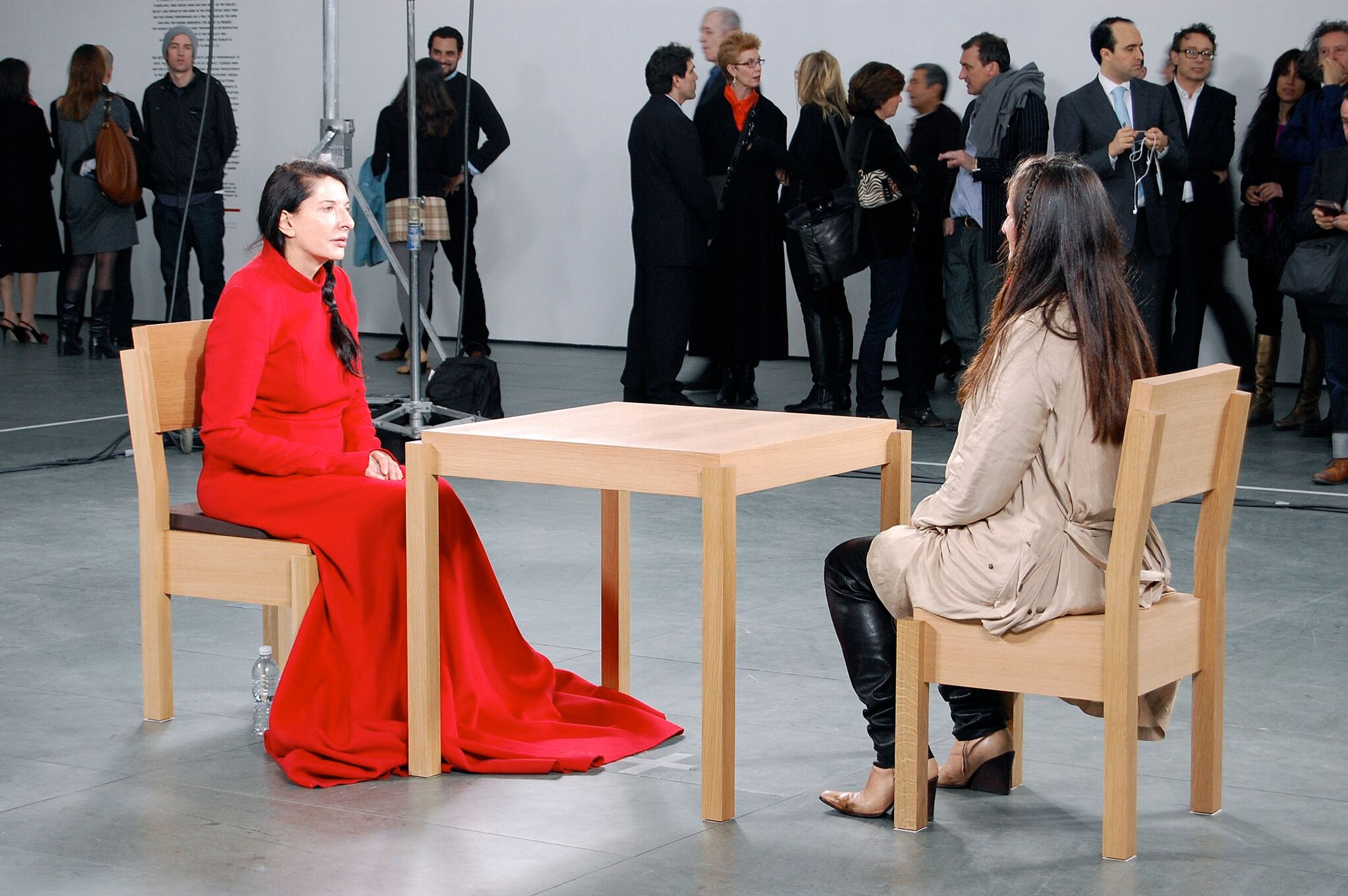 People watching a woman in a red speaking at a desk to a woman in a beige coat