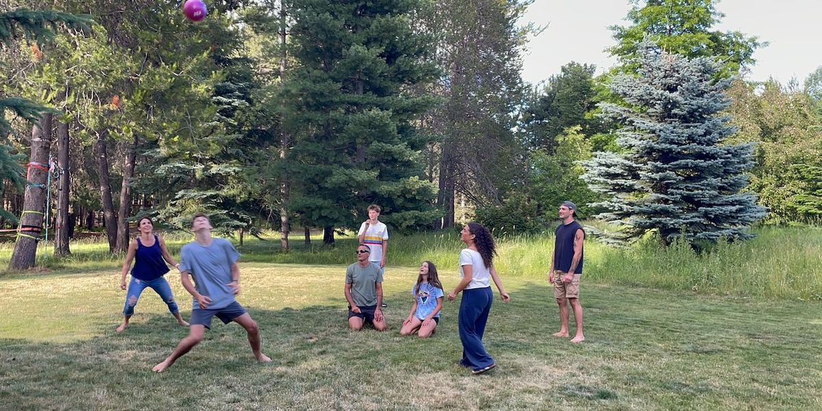 Family playing an outdoor game with a volleyball with a couple sitting in the middle and the rest standing in a circle.