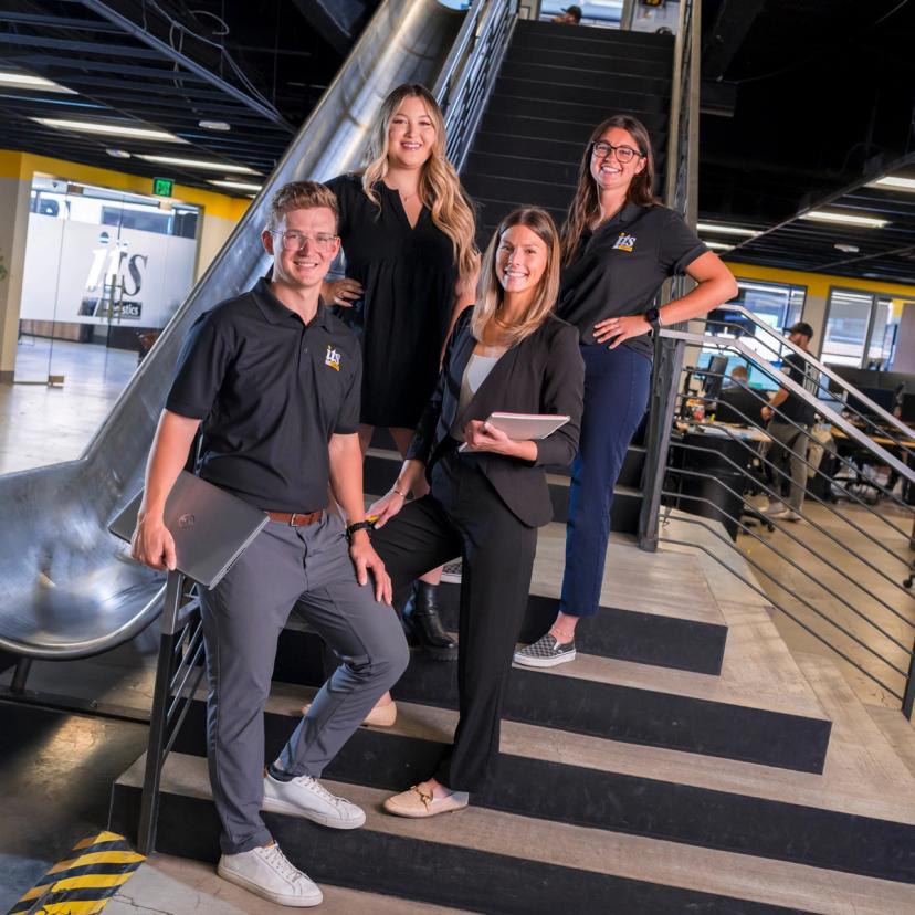 Four ITS Logistics team members posing together with a slide and stairs in the background