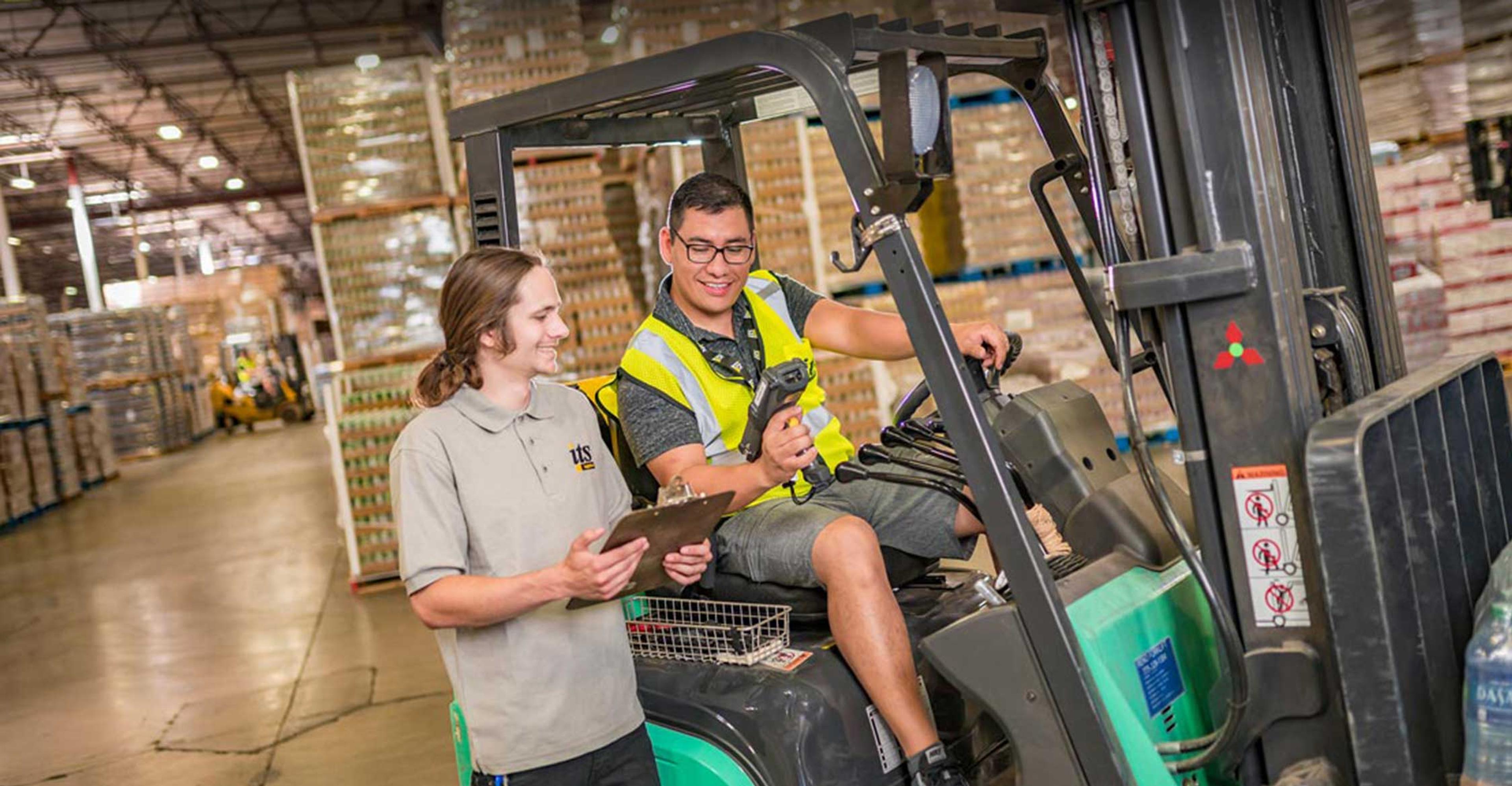 2 men in a warehouse, one on a forklift, the other standing by it.