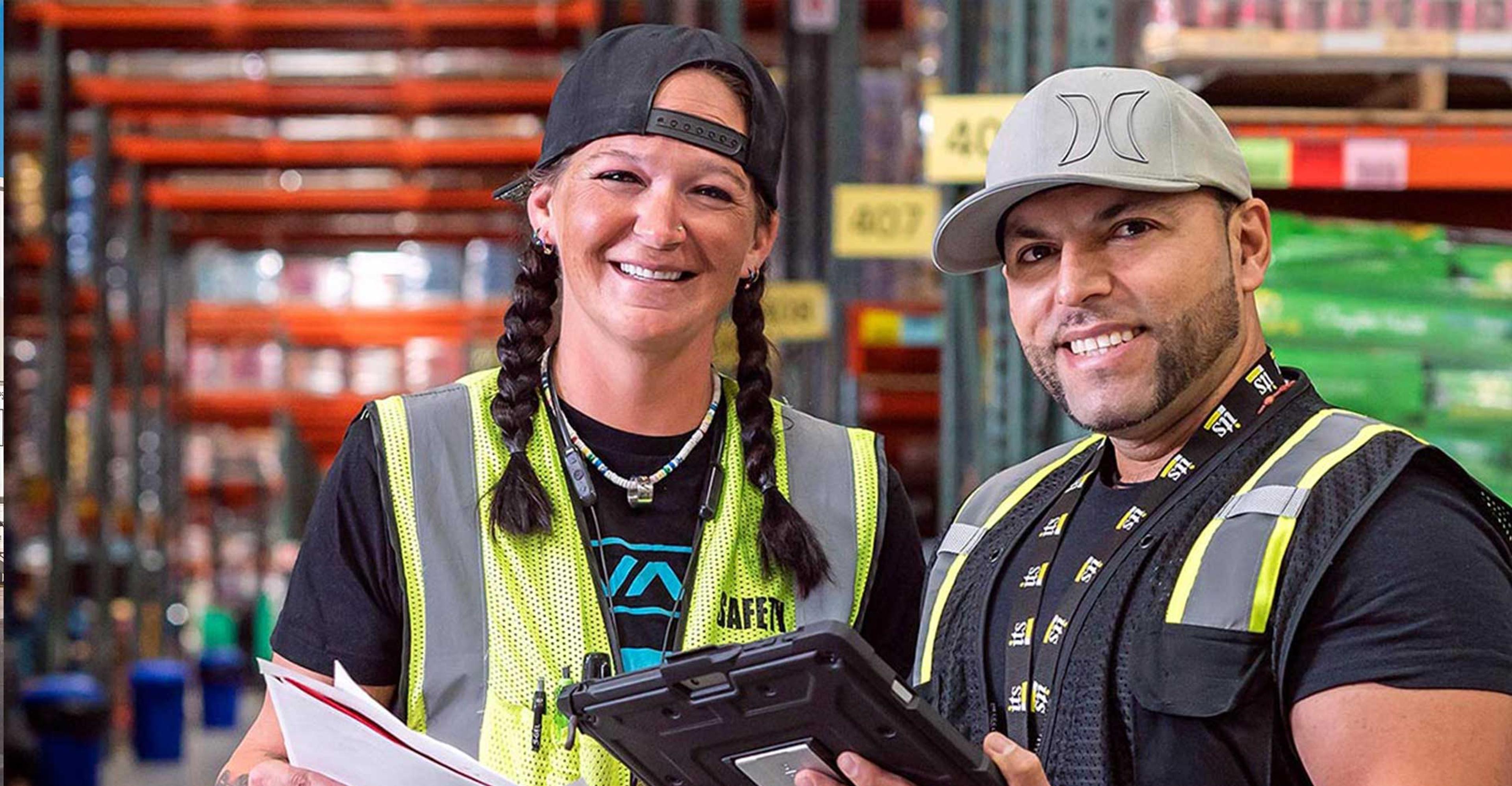 2 ITS warehouse employees smile at the camera while holding papers and an iPad.