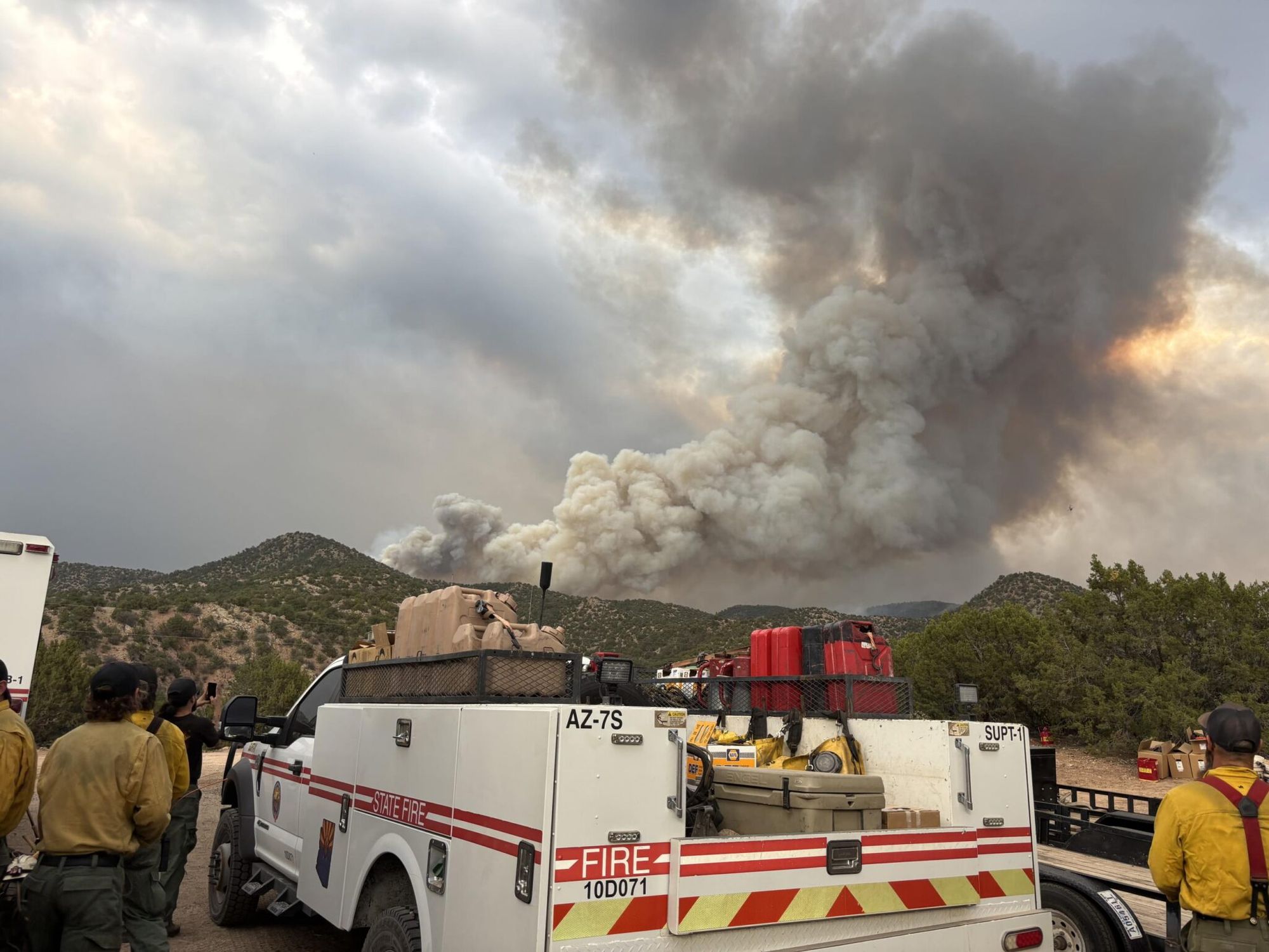 A large smoke plume from the Frijoles Canyon Fire rises over the mountains as firefighters and a state fire vehicle stand by in the foreground