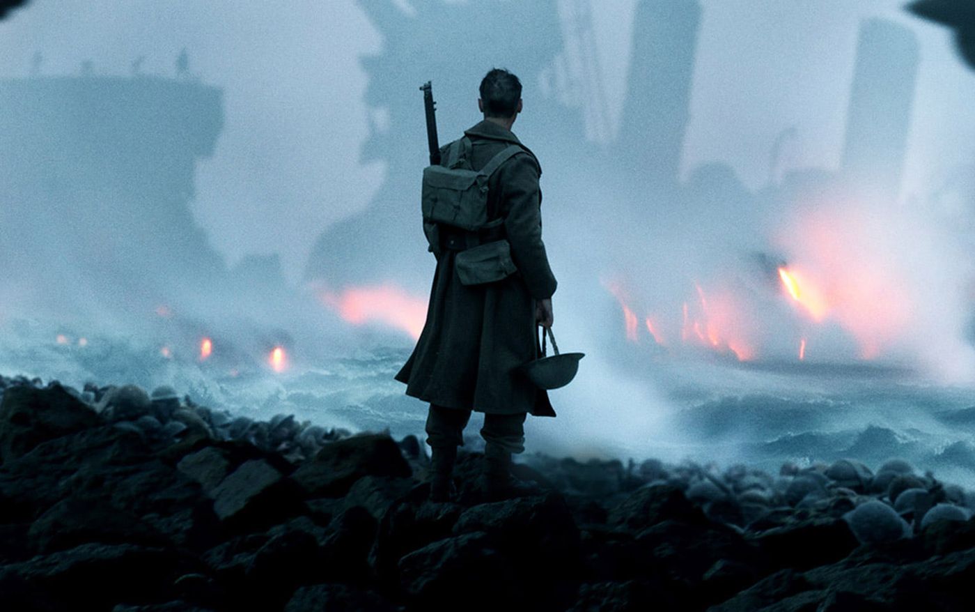 Still from Dunkirk: a soldier with a rifle stands on a rocky beach facing smoke and fire