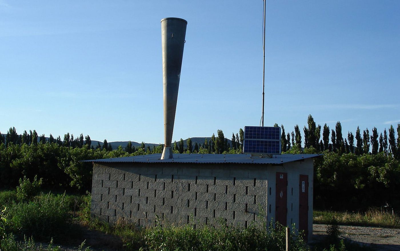 Hail cannon with a tall metal horn on top of a concrete block building in a field