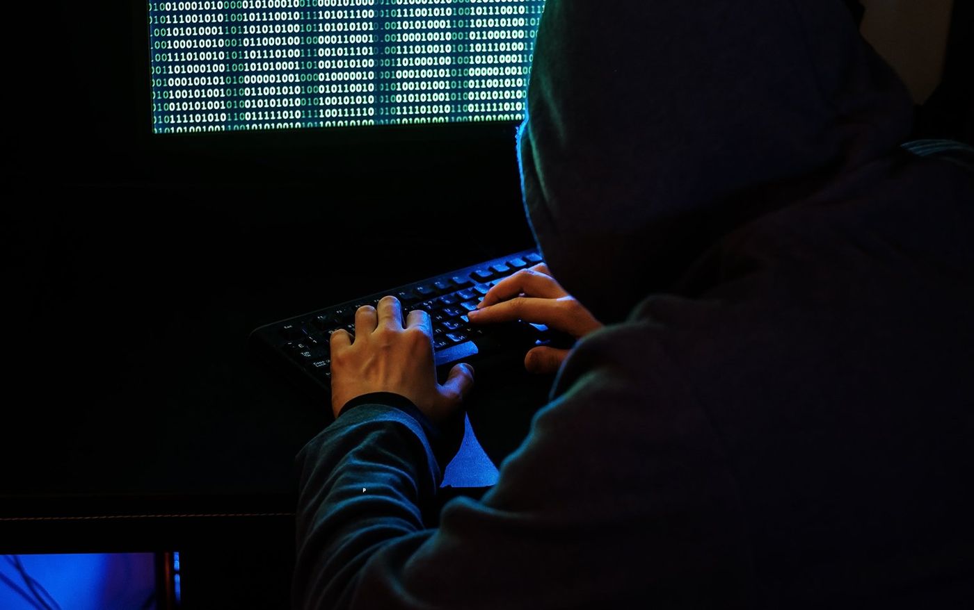 Hooded figure typing at a keyboard in front of a screen of binary code, illustrating video piracy