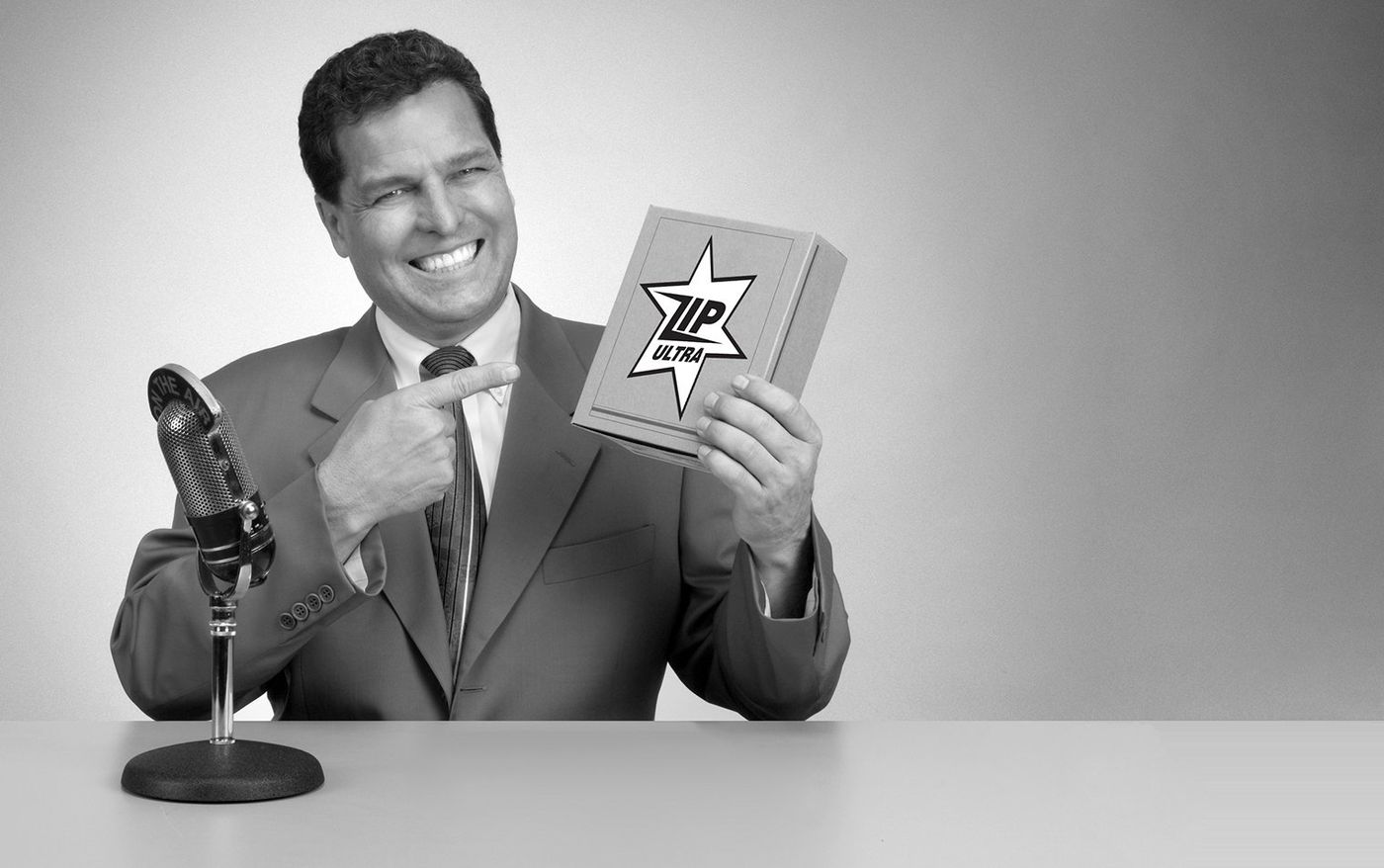 Black and white photo of a grinning TV pitchman pointing at a product box beside a vintage microphone