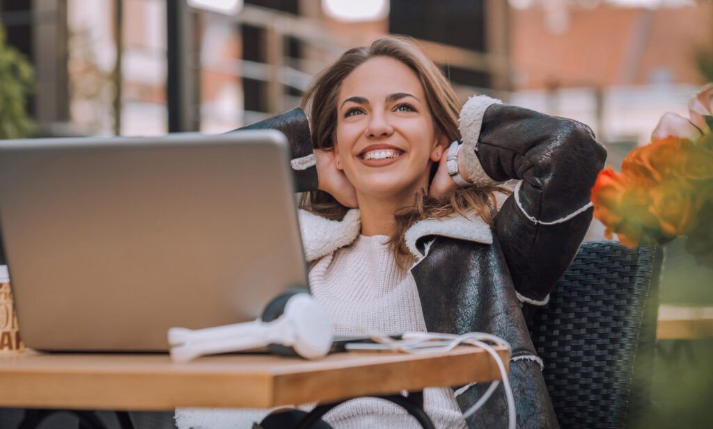 Smiling woman leaning back at her laptop at an outdoor cafe table, working remotely