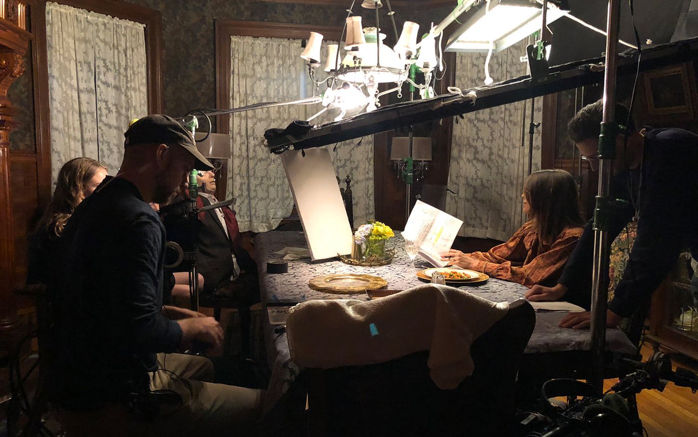 Behind the scenes on the Father Figurine shoot: actors at a dinner table lit by overhead lights while crew work around them