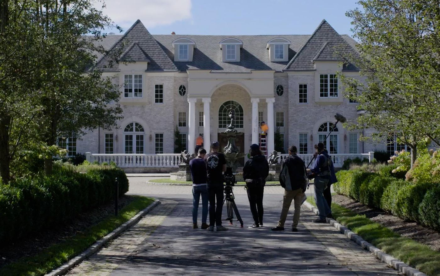 Film crew with camera and boom mic on the driveway of a large mansion during the shoot