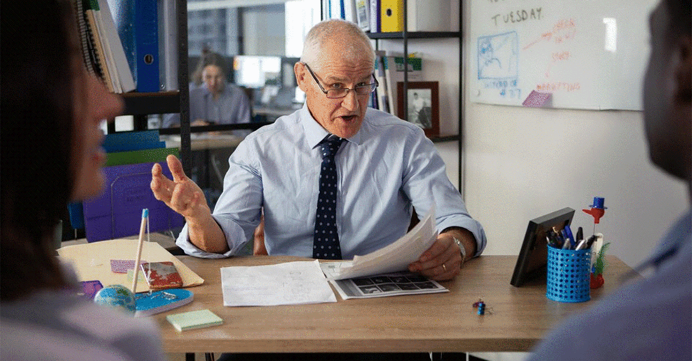 Older executive in a shirt and tie reviewing papers and gesturing during an office meeting