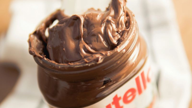 Un barattolo di Nutella aperto da cui è stato estratto un cucchiaino, rivelando la sua morbida e cremosa crema al cioccolato e nocciole. Il barattolo è adagiato su un panno bianco con sfondo sfocato.