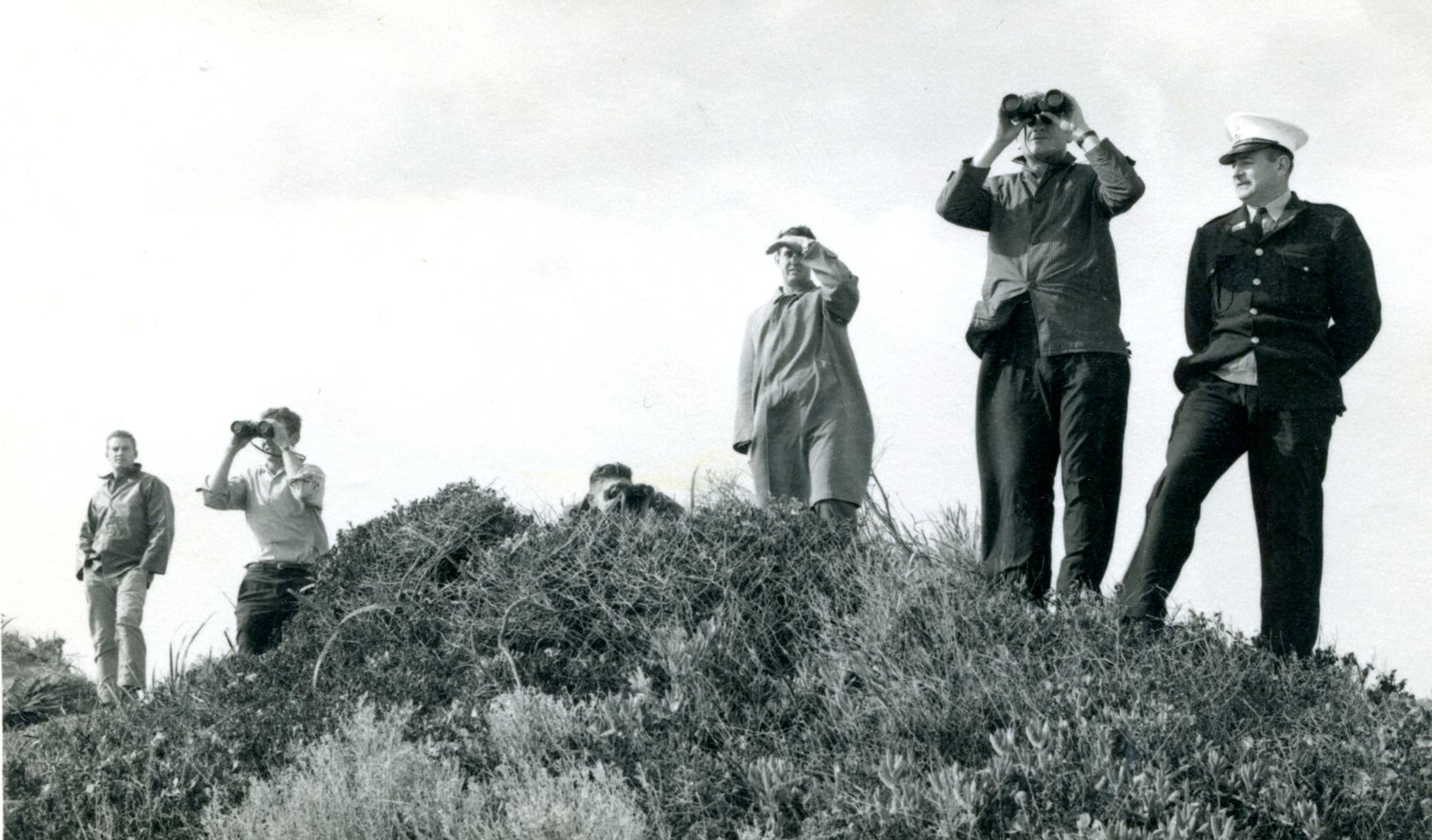 Black and white photo of men on sand dune looking for Avaneta