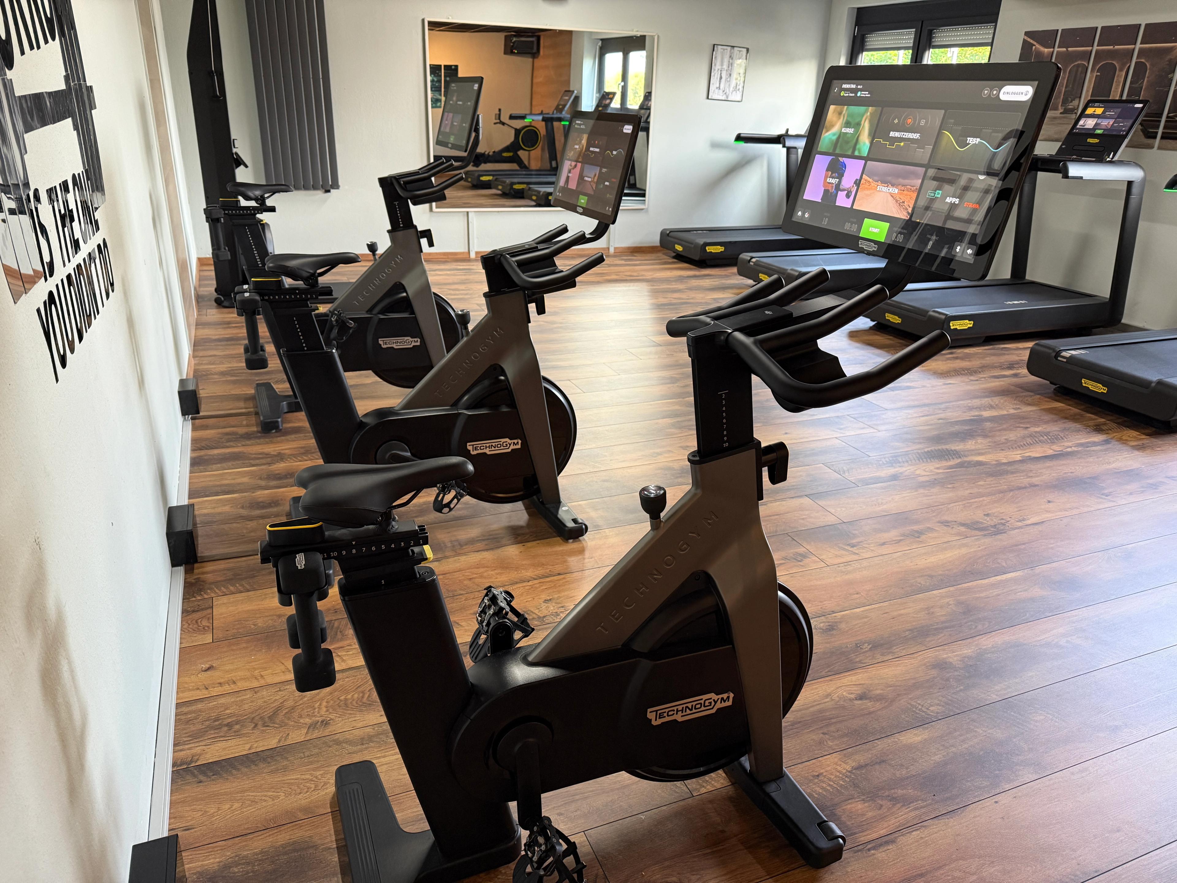 Ein Fitnessstudio mit mehreren schwarzen Technogym Indoor-Cycling Rädern, jedes ausgestattet mit großem Touchscreen-Display