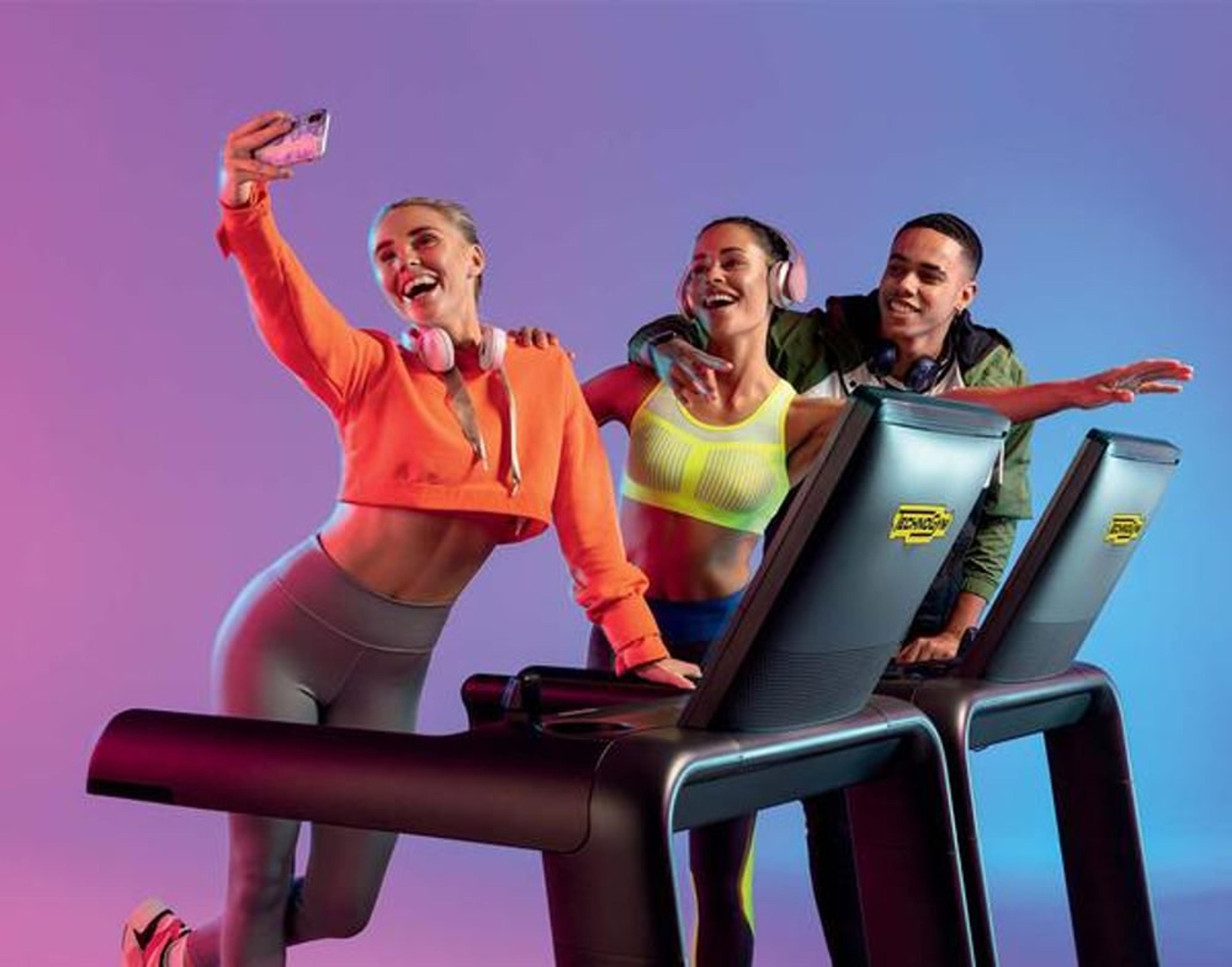 Drei junge Erwachsene auf Laufbändern, die ein Selfie machen, mit bunten Sportoutfits und Kopfhörern