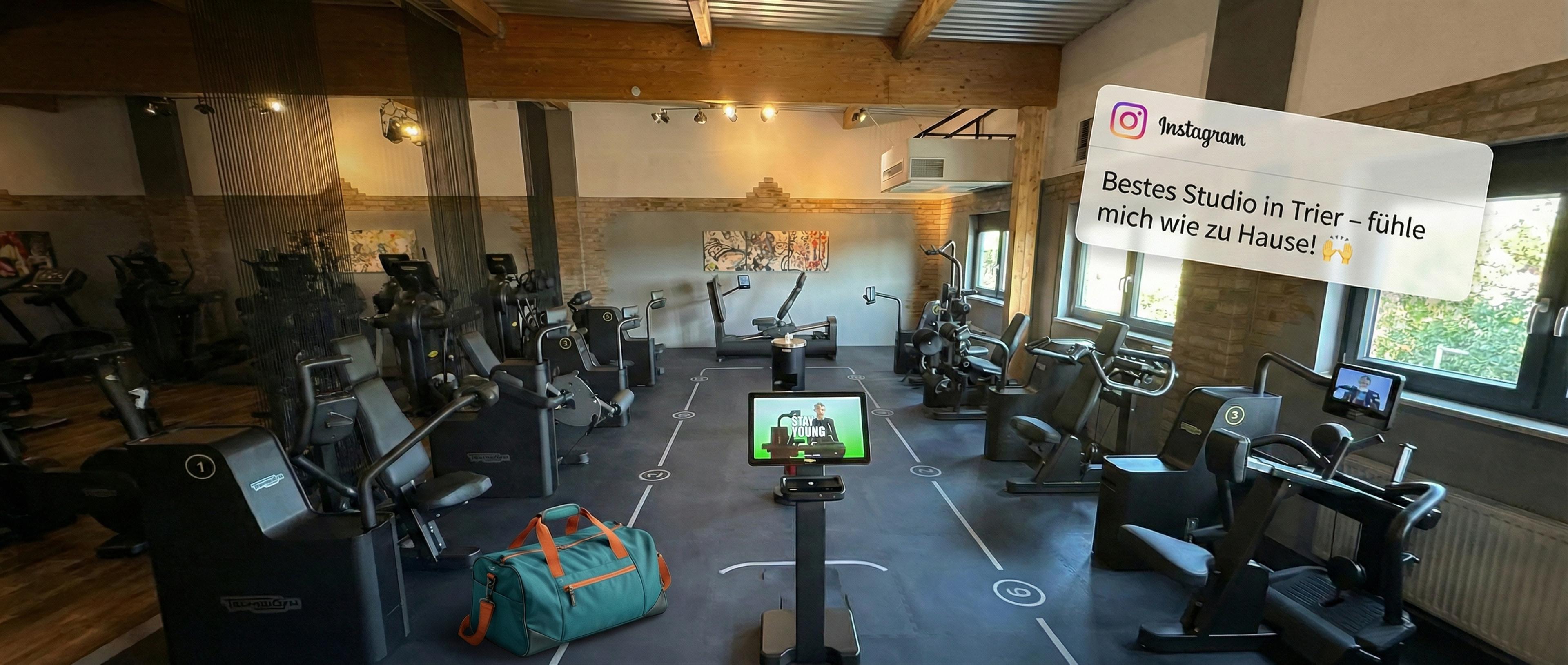 Ein heller Trainingsraum mit mehreren schwarzen Technogym-Kraftgeräten, sichtbaren Holzbalken und großen Fenstern