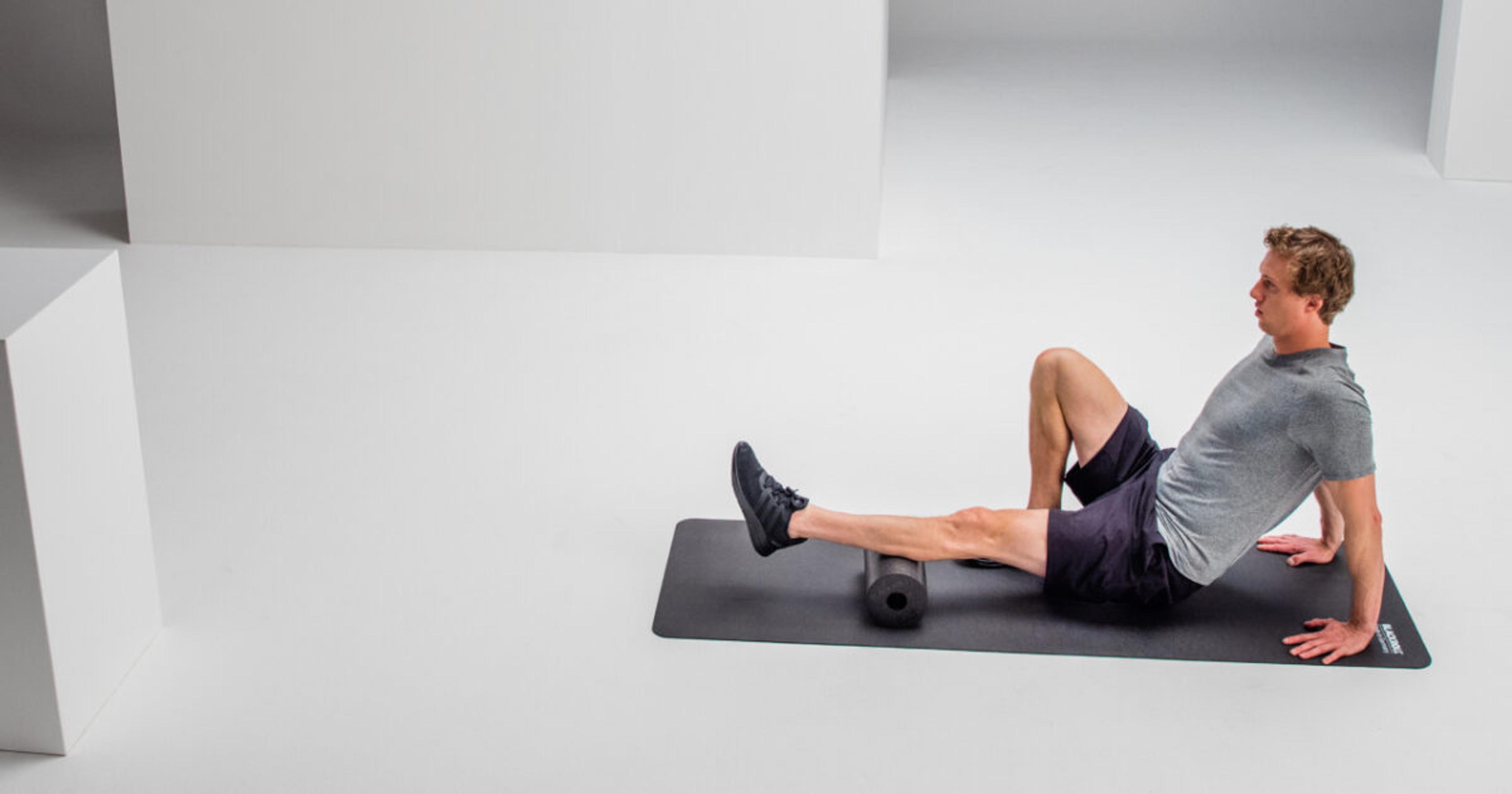 Ein Mann massiert seine Wade mit einer schwarzen Schaumstoffrolle auf einer dunklen Trainingsmatte in einem weißen Studio